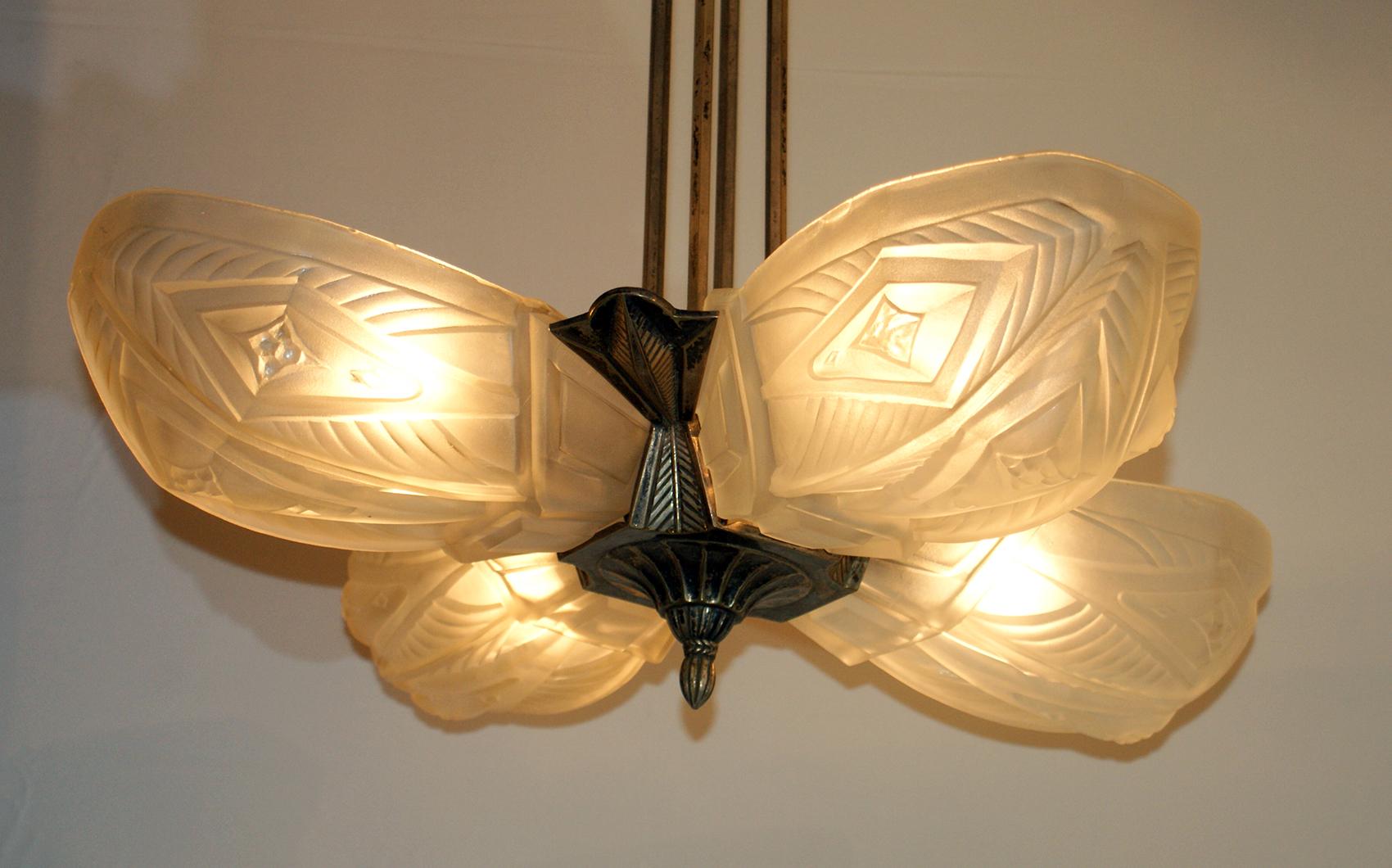 A stunning Art Deco Chandelier crafted in France and signed Francis Hubens.
This chandelier features four frosted glass panels with ornate detail work, held by a matching nickeled bronze hardware.
Having four candelabra sockets for the glass