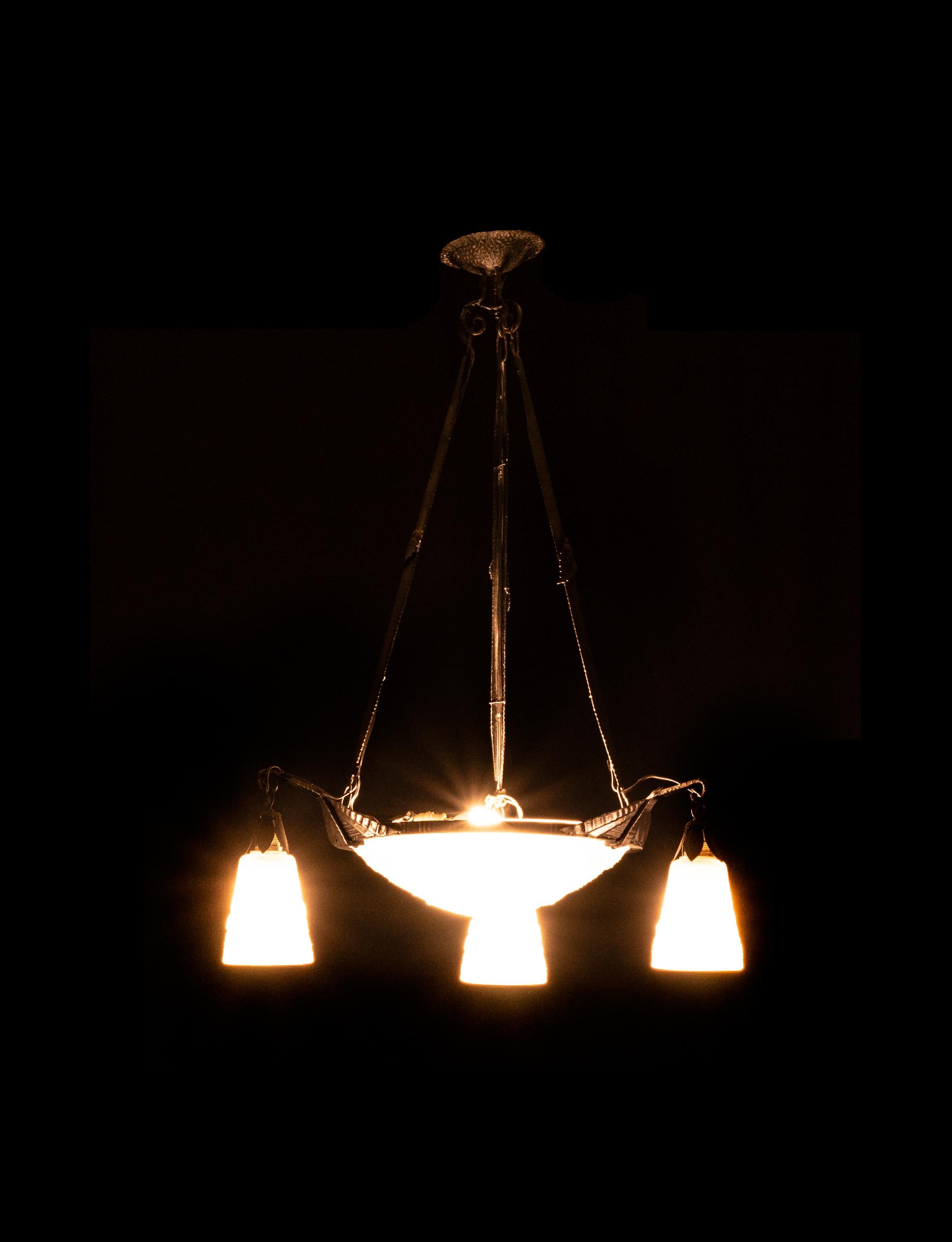 Lampadario francese Art Deco degli anni '20, realizzato da Muller Frères a Lunéville. 
Il lampadario presenta una struttura in ferro battuto scuro che sostiene una ciotola centrale in vetro smerigliato con tre paralumi a forma di tulipano. 
Ogni