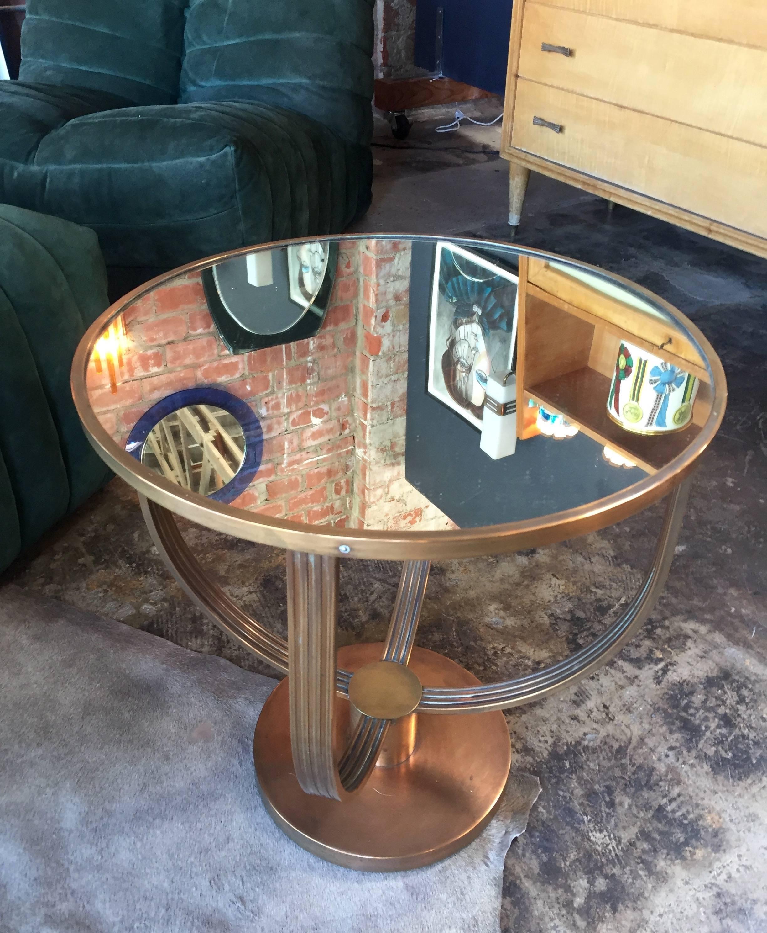 This smaller size cocktail table attributed to Jean Michel Frank has a very elegant Art Deco design: a round coffee table in slightly darkened brass. Four curved pieces separate the base and top. The top is fitted with a mirrored glass insert.