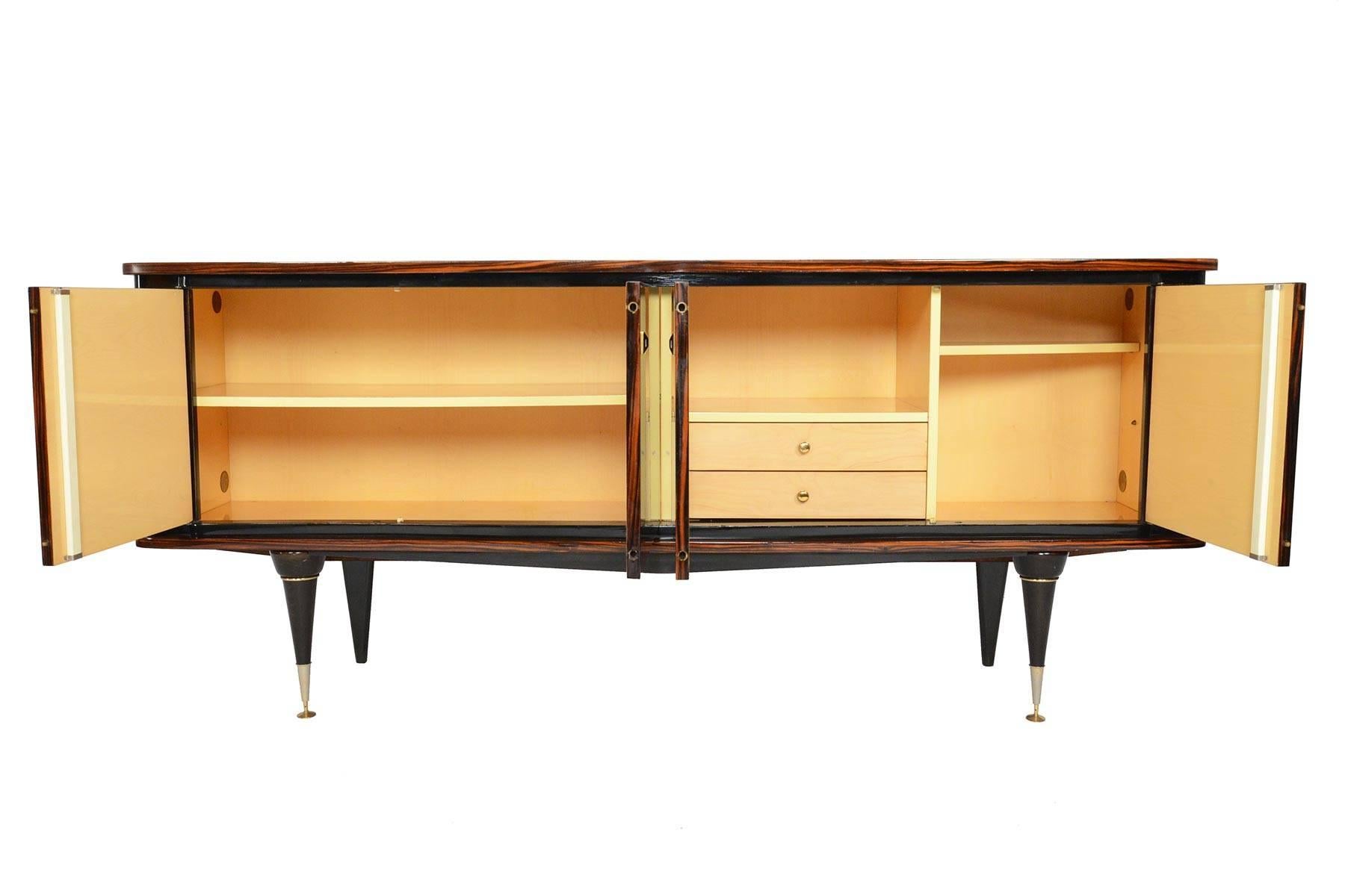 This French Art Deco credenza is cased in exotic Macassar and features rounded corners and ornate ebony legs. Beautiful book match veneer accentuates its soft shape. Two pairs of locked doors open to reveal three bays. The spacious left bay offers