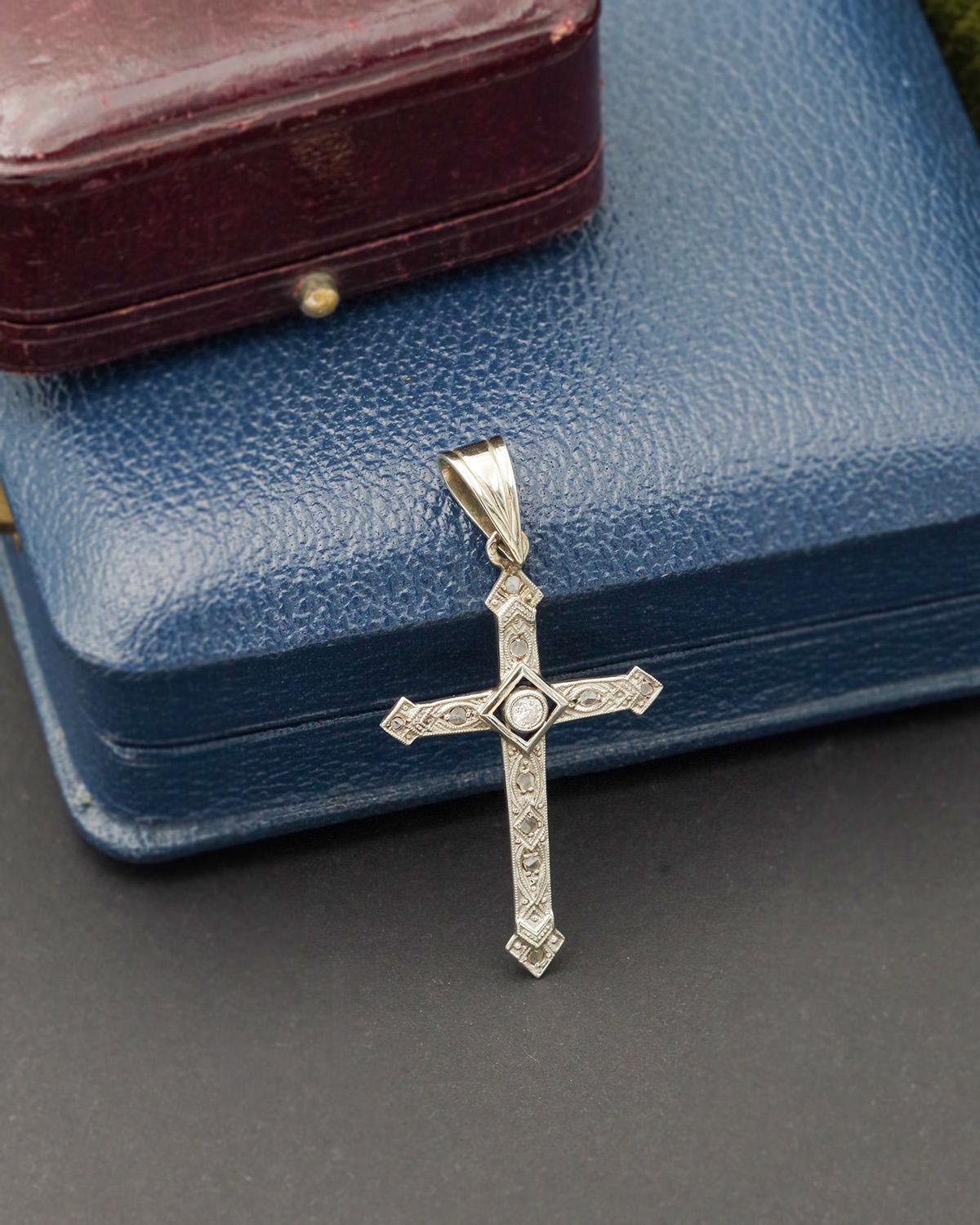 Art Deco diamond cross in 18 karat white gold. Antique cross pendant from the early 20th century set with a 0.07 ct old-cut diamond. The arms of the cross are set with ten flat diamonds, highlighted by a delicate arabesque and beaded frieze design.