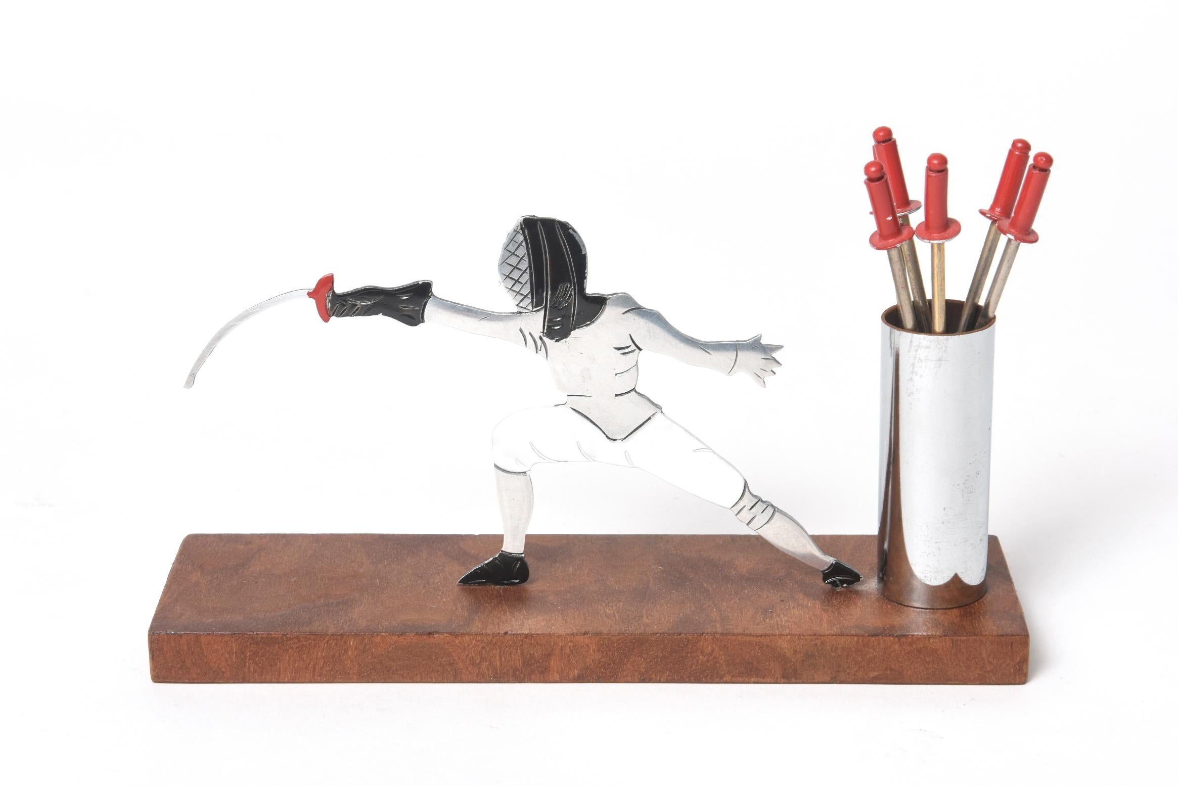 A French Art Deco chrome cocktail pick set featuring a fencer lunging with his sword in hand. The six epee sword picks fan out in a chrome cylinder stand. The picks have painted red finials. These cocktail pick holders (