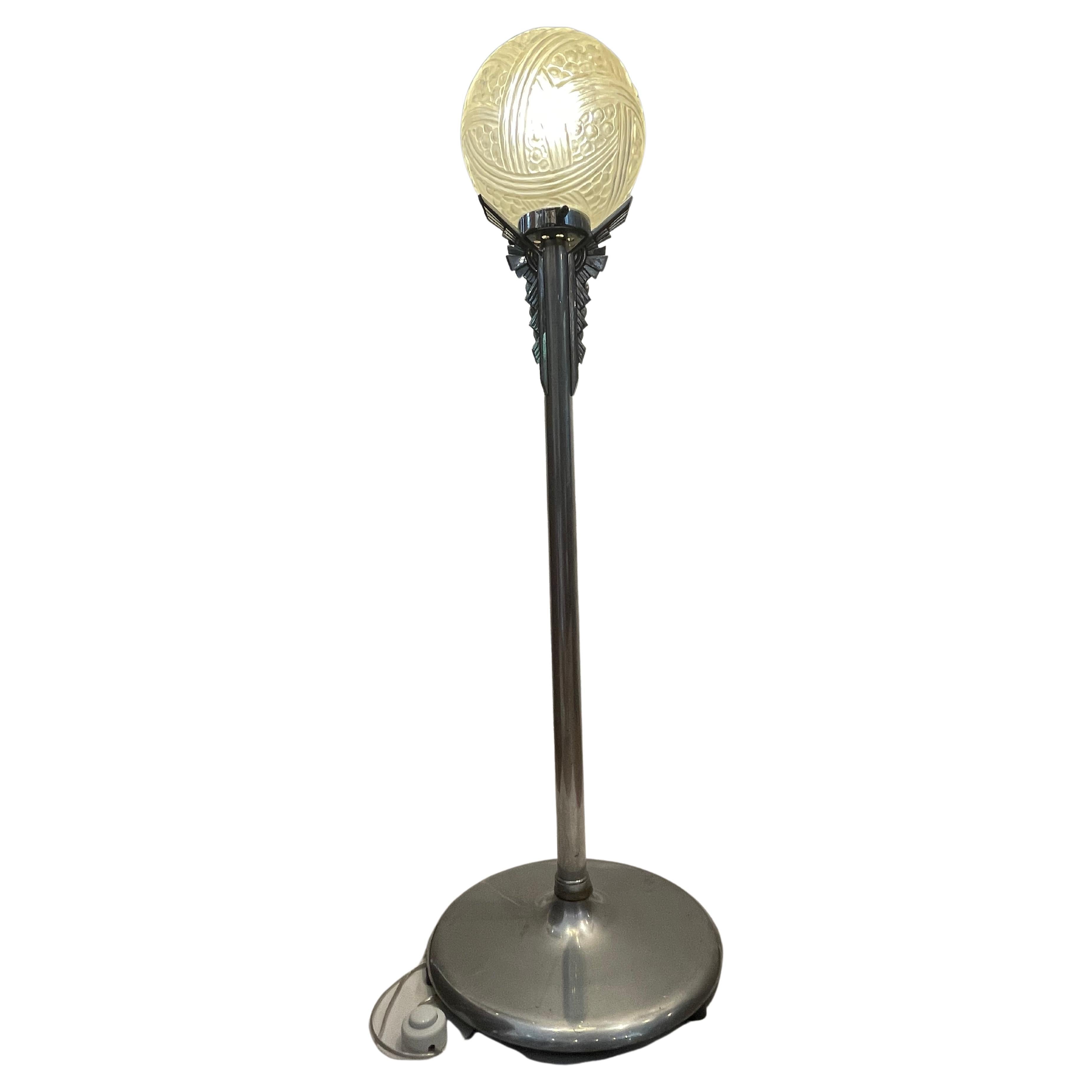Lampada Art déco con base cromata. La colonna è sormontata da una decorazione geometrica a punto su tre dimensioni, il tutto poggia su una base ampia e stabile.
La sfera pressata in stampo di vetro opaco e satinato dà un effetto di luce soffusa