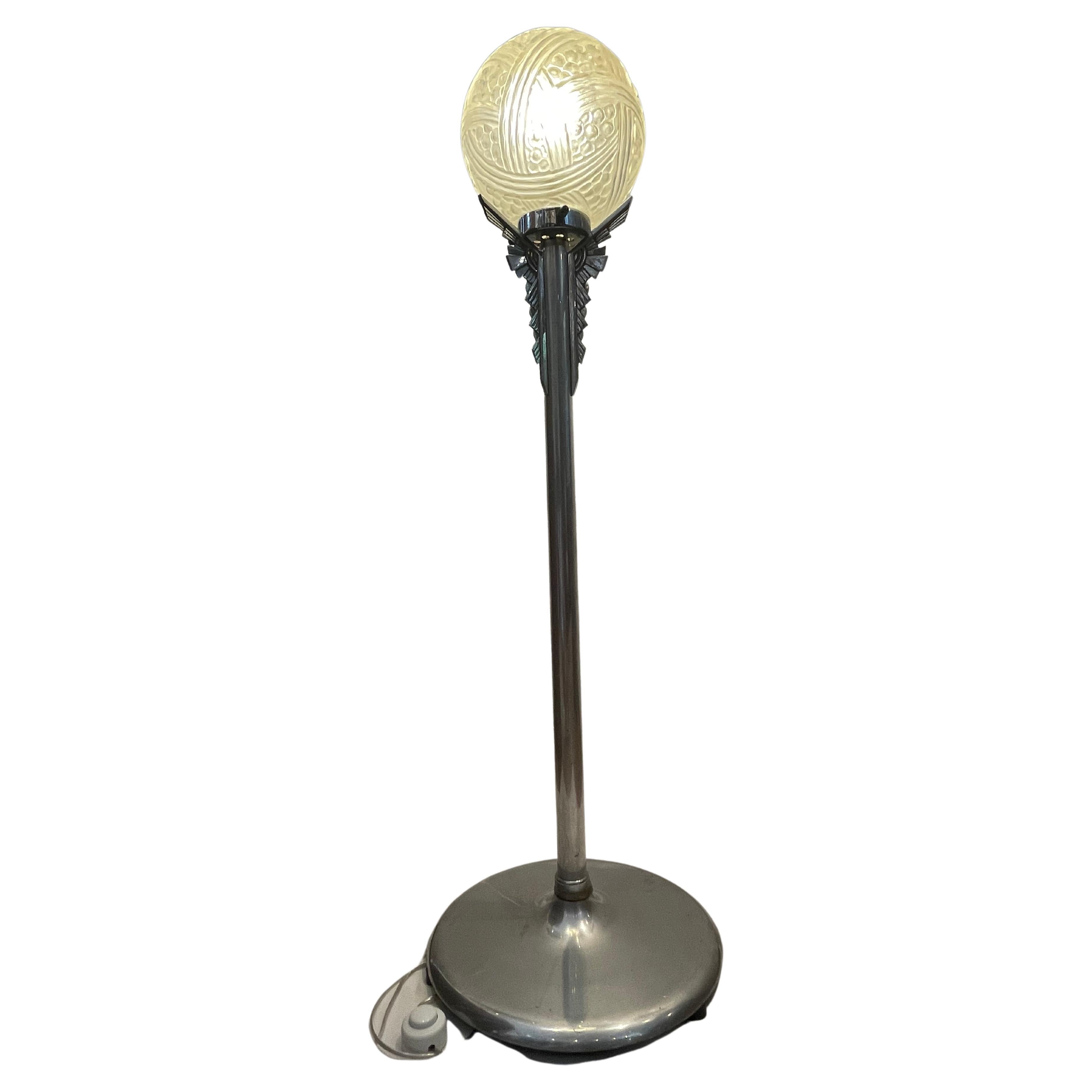 Lampe Art déco avec base chromée. La colonne est coiffée d'un décor géométrique en pointe sur trois dimensions, le tout reposant sur une base large et stable.
La boule pressée en verre moulé mat et satiné un décor stylisé donne un effet de lumière