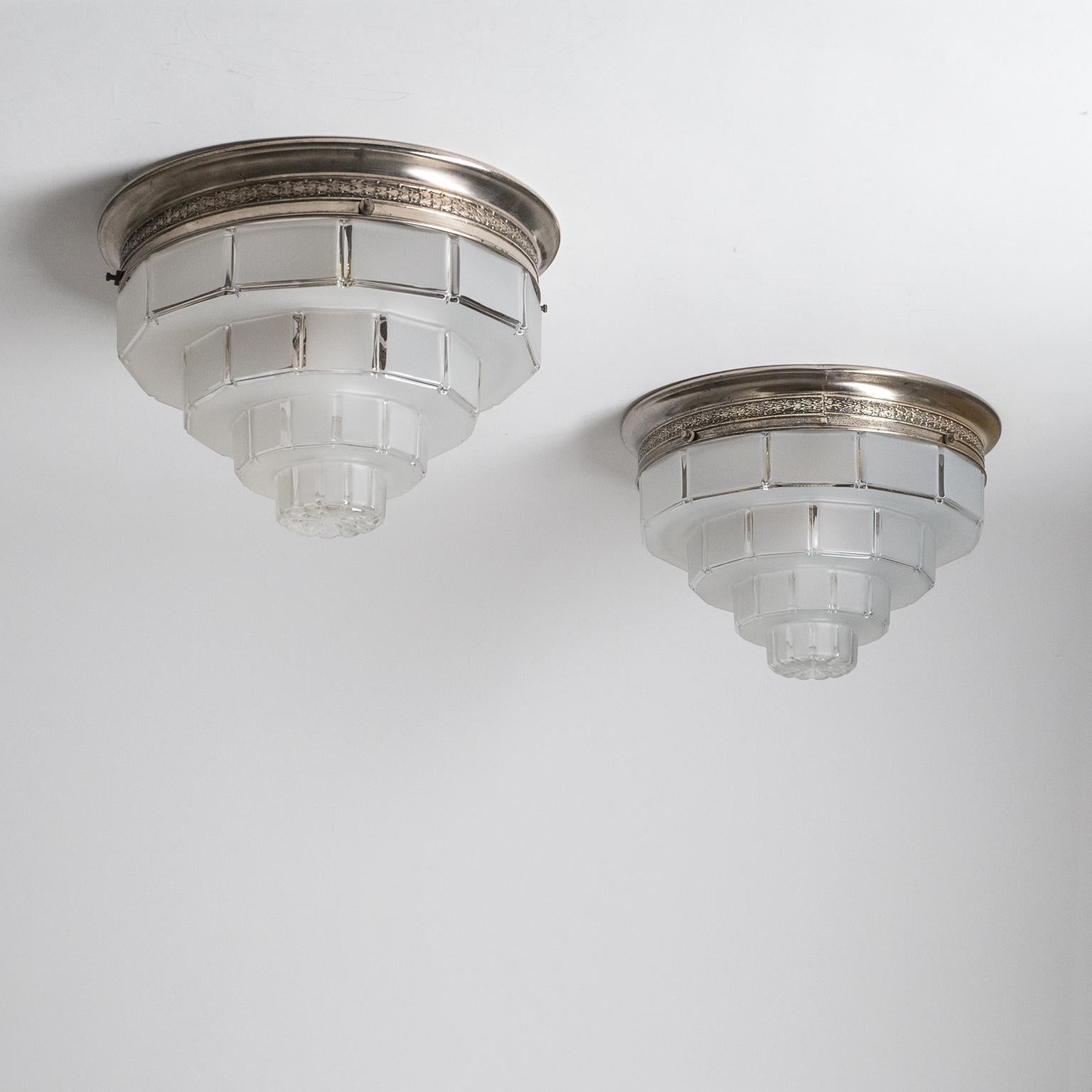 Pair of French Art Deco flush mounts, circa 1930. An intricately detailed nickeled brass backplate holds a multi-tiered 'skyscraper' glass diffuser which is frosted on the outside with clear glass accents. Good original condition with some