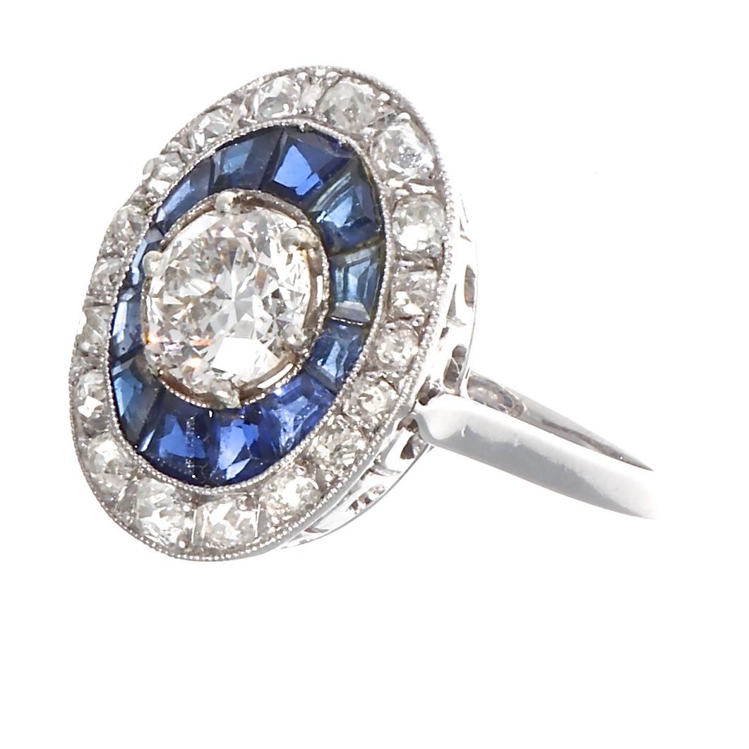 This thrilling ring features a GIA certified 1.05 carat, F color VS1 clarity     old European cut diamond as the point of focus. Creating even more luminosity are the concentric rows of 12 emerald cut sapphires and 18 old European cut diamonds. The