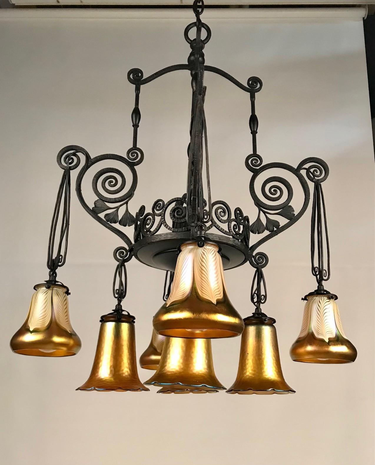 This elegant chandelier combines the vigor of hand-hammered wrought iron with the stylish iridescence of Quezal shades. The scrolling and leafy frame hangs from its original chain and canopy and supports four Aurene shades beneath four Pulled