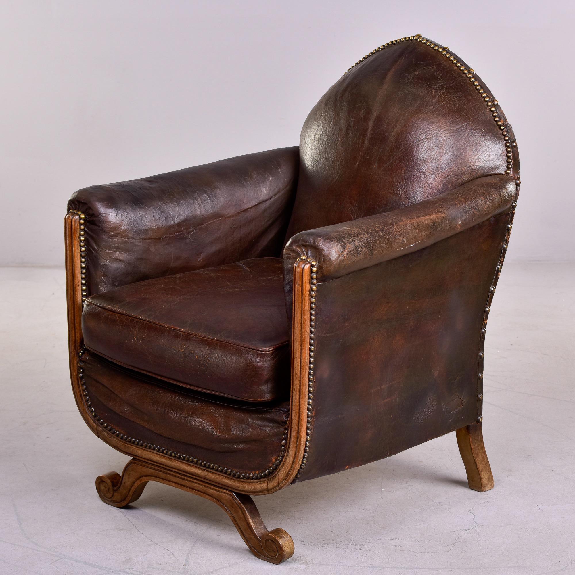 Circa 1930s French leather club chair with wood frame. Camel back chair with rolled arms, exposed wood frame, nail head trim and curved, scrolled front leg. Original dark brown leather upholstery shows visible wear throughout. Chair is structurally