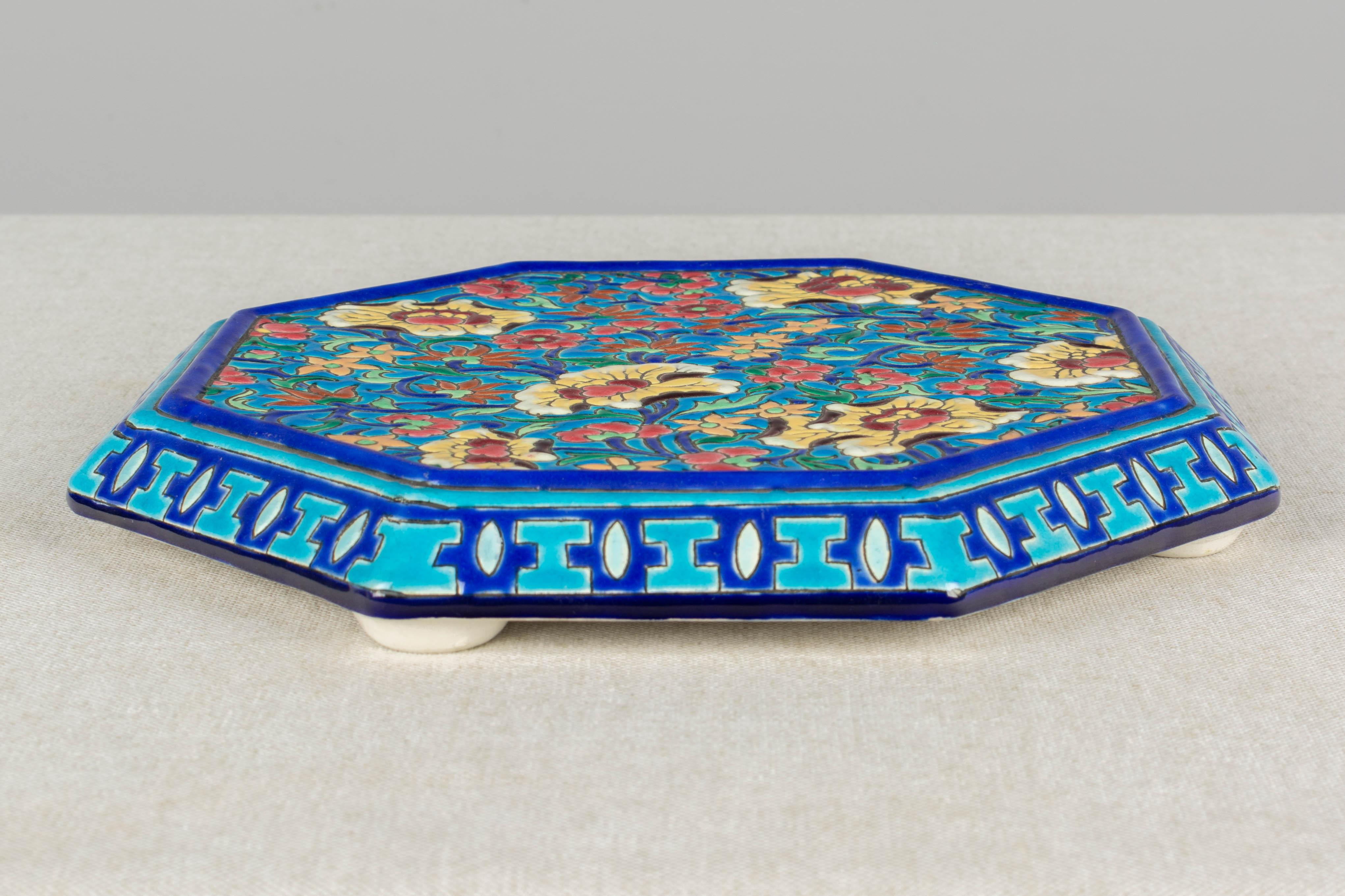 A French Art Deco Longwy octagonal shaped hand painted ceramic trivet decorated in the style of chinoiserie cloisonné enamel. Bright turquoise blue ground with a colorful overall floral design in yellow, green, orange and pink. Impressed makers mark