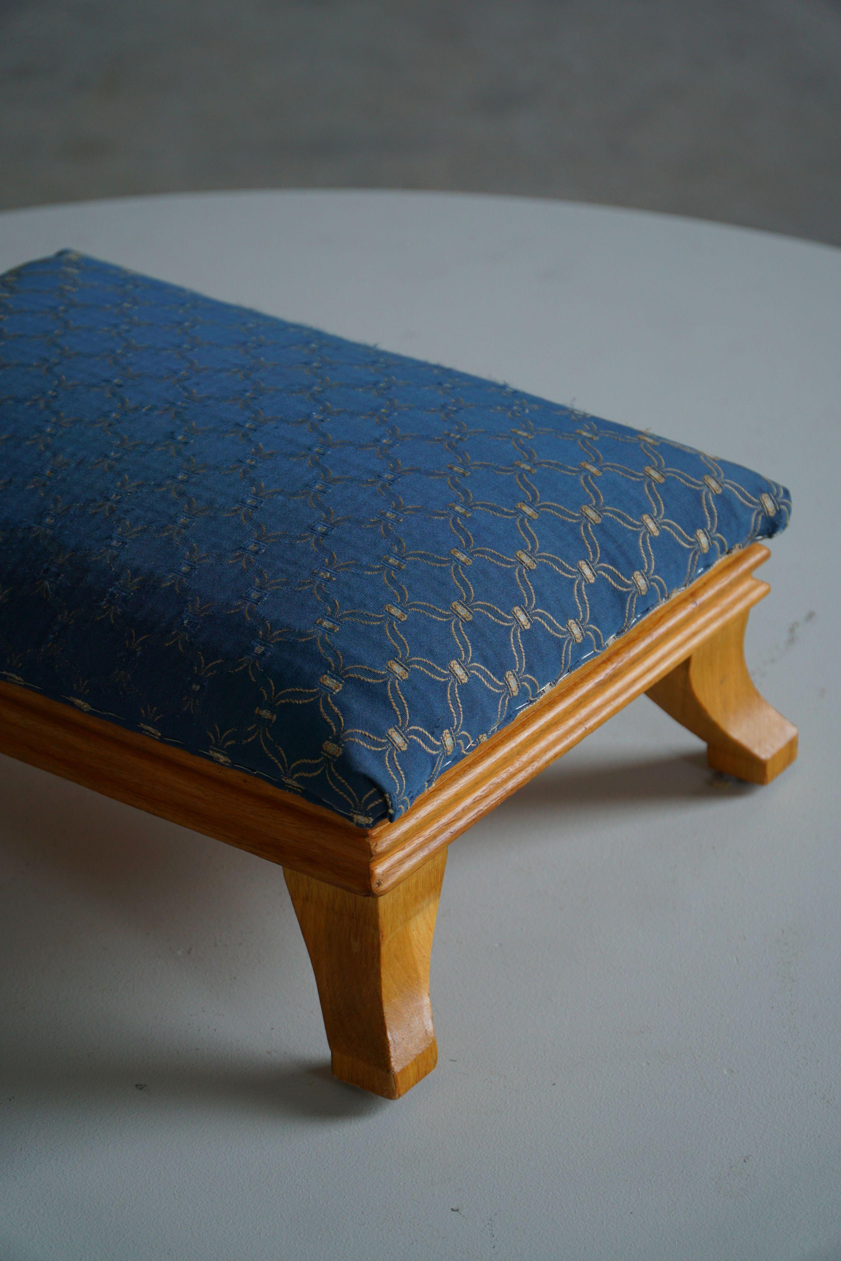 Un affascinante sgabello basso in stile Art Déco dalla forma rettangolare. Fabbricato negli anni '40 da un ebanista francese, il pezzo è realizzato in legno di olmo e rivestito in un tessuto blu vintage ornato da un elegante motivo dorato, che