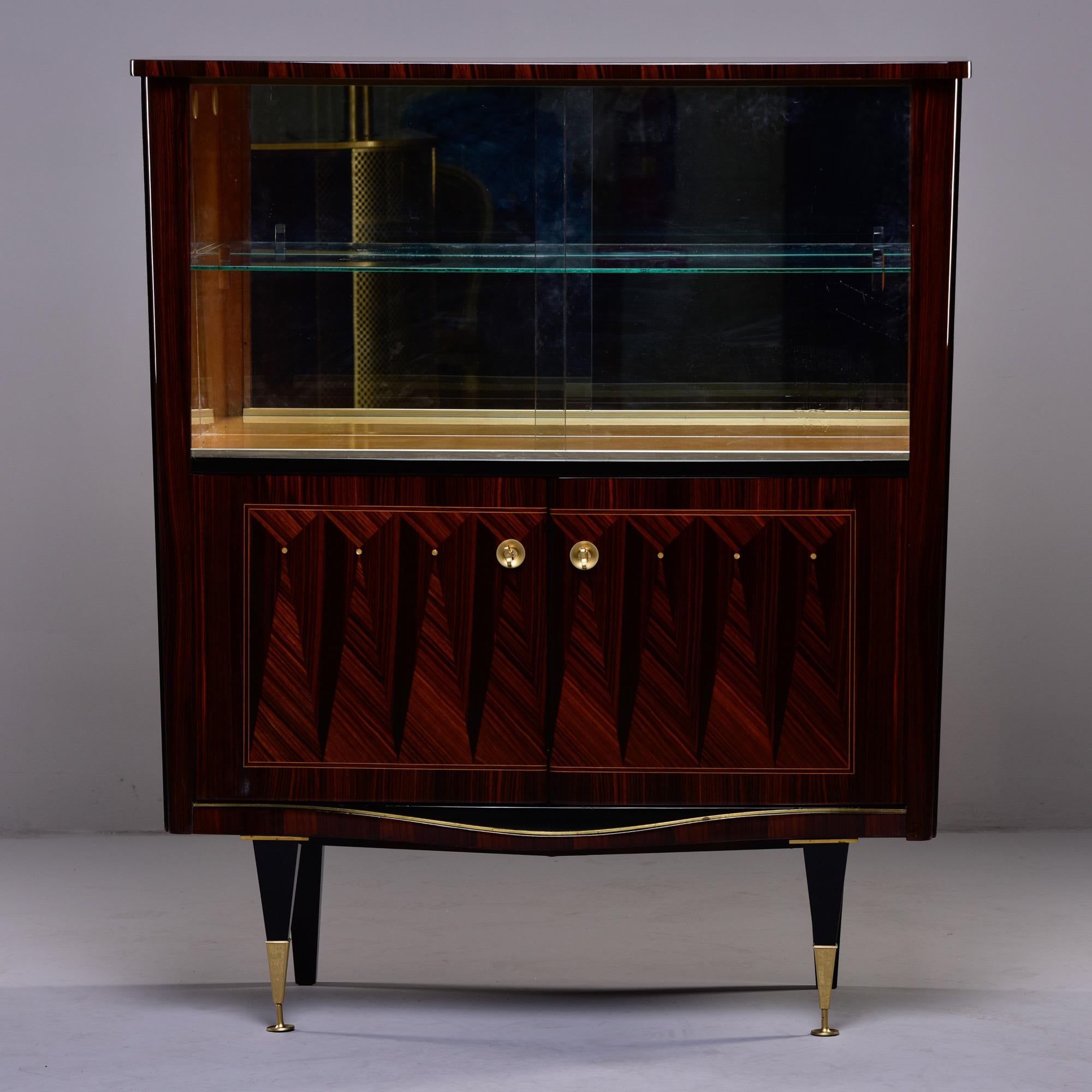 Circa 1940s French Art Deco bar cabinet or vitrine in macassar with marquetry pattern on lower cabinet doors, contrasting maple interior trim and single adjustable glass shelf. Brass capped front legs. Top compartment has sliding glass doors and