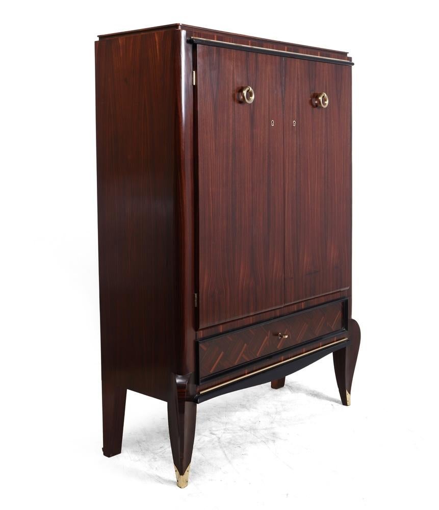 This Art Deco Macassar cabinet has two doors with adjustable shelves behind and a single lockable drawer below. The cabinet stands on short Ruhlman style saber legs with brass tips.