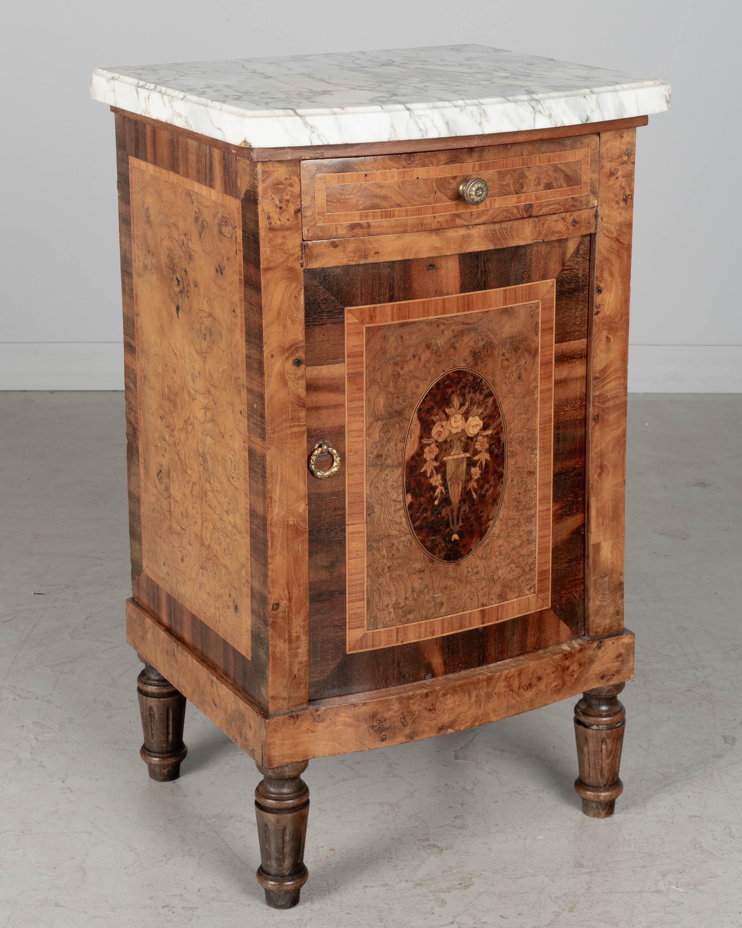 Ein französischer Nachttisch im Art Déco-Stil mit Intarsien aus Ulme, Ahorn und Nussbaum. Schublade mit Schwalbenschwanz über einer Schranktür mit ovalem Intarsienmedaillon mit Blumenurne. Beschläge aus Messingguss. Gedrechselte, konische Beine. Die