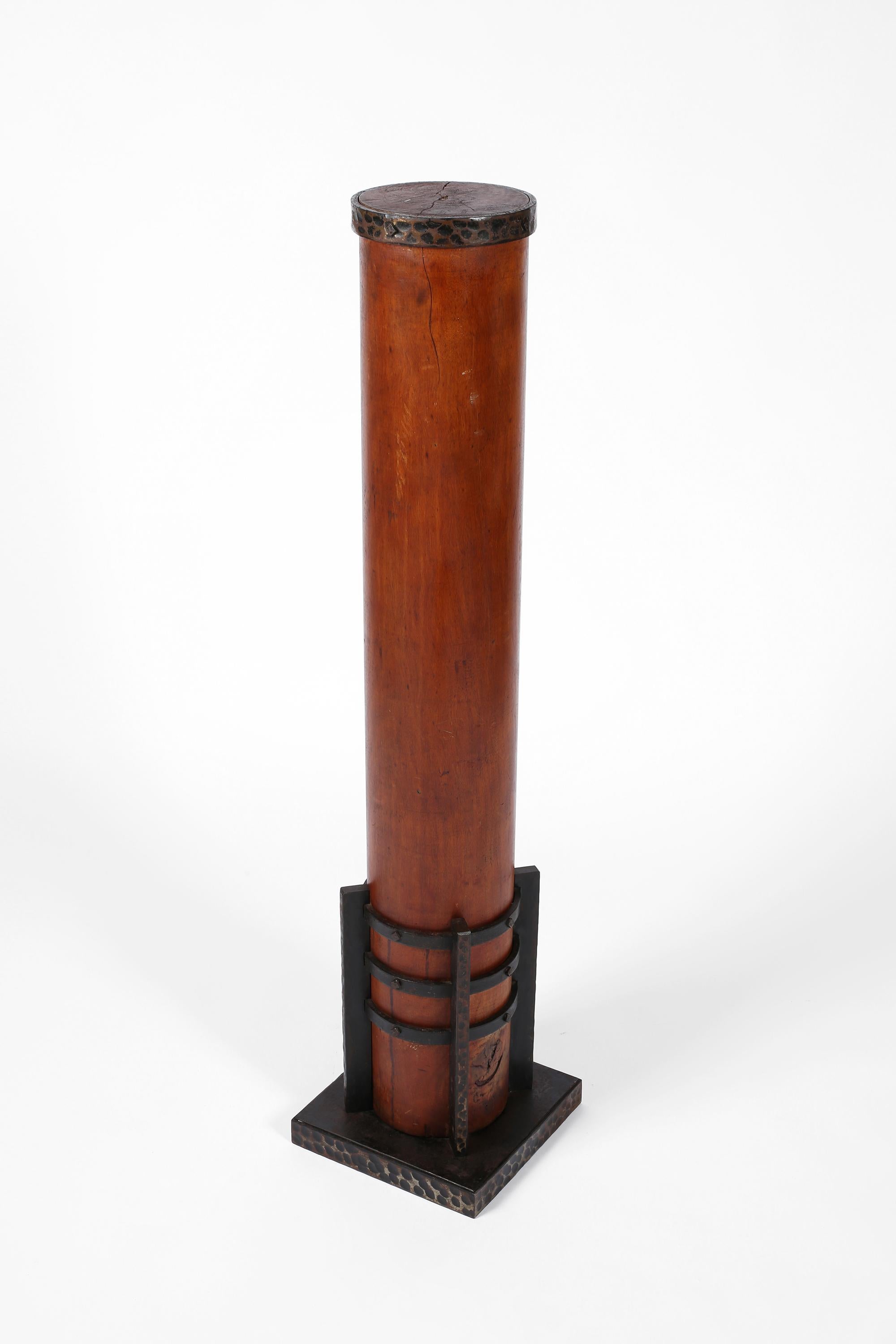 Un imponente basamento a colonna in mogano e ferro forgiato di Michel Zadounaïsky (1903-1983). Il cilindro di legno massiccio intagliato è contenuto nel supporto in ferro, decorato con la caratteristica finitura martellata di Zadounaïsky, impressa