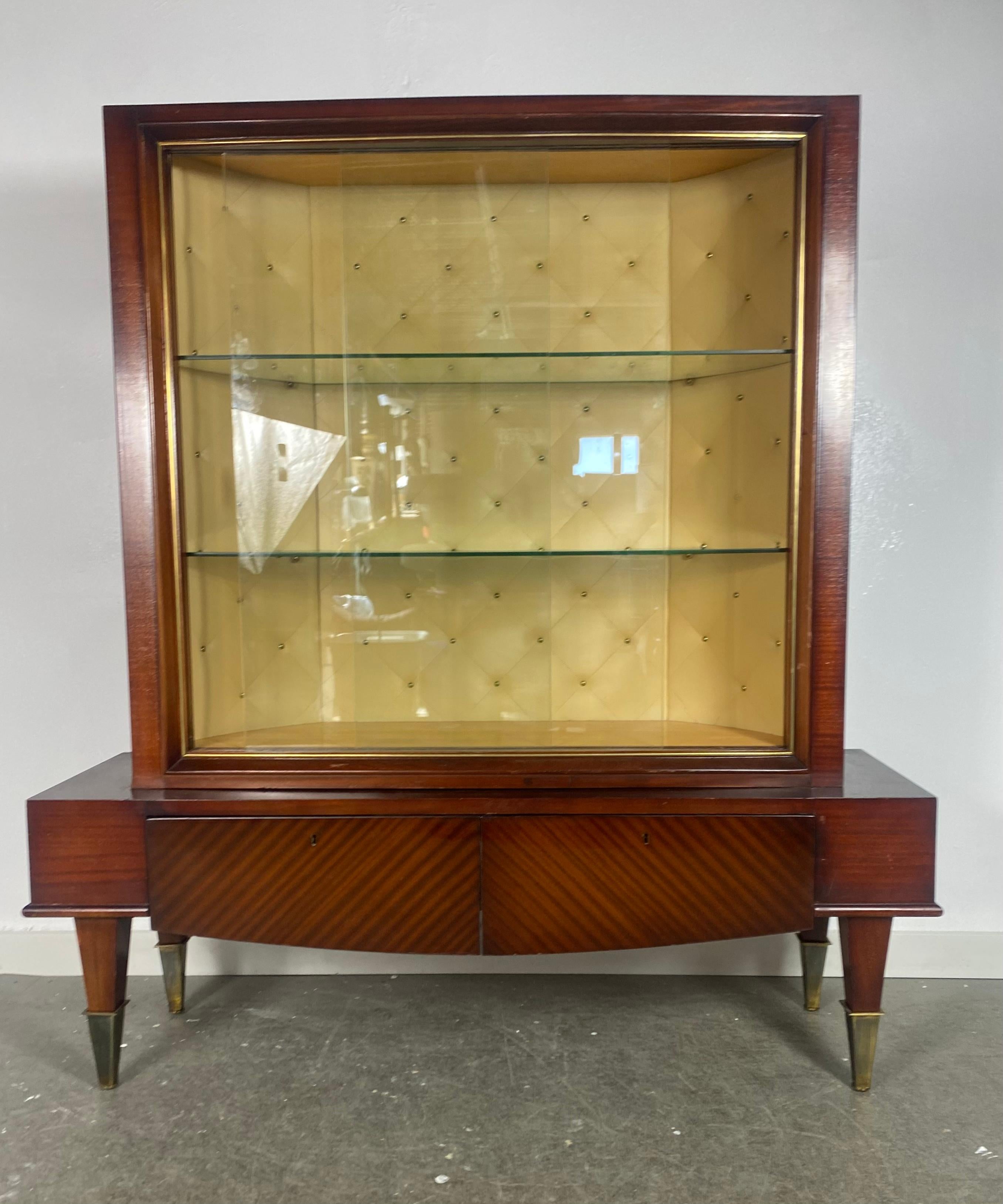 Stunning French Art Deco / Moderne' vitrine, bar, cabinet attrib. to Jules Leleu 1940s. Classic design, wonderful grained cherry mahogany wood. Button-tufted interior, two glass shelves, sliding doors. Two lower drawers. Nice brass cap accents to