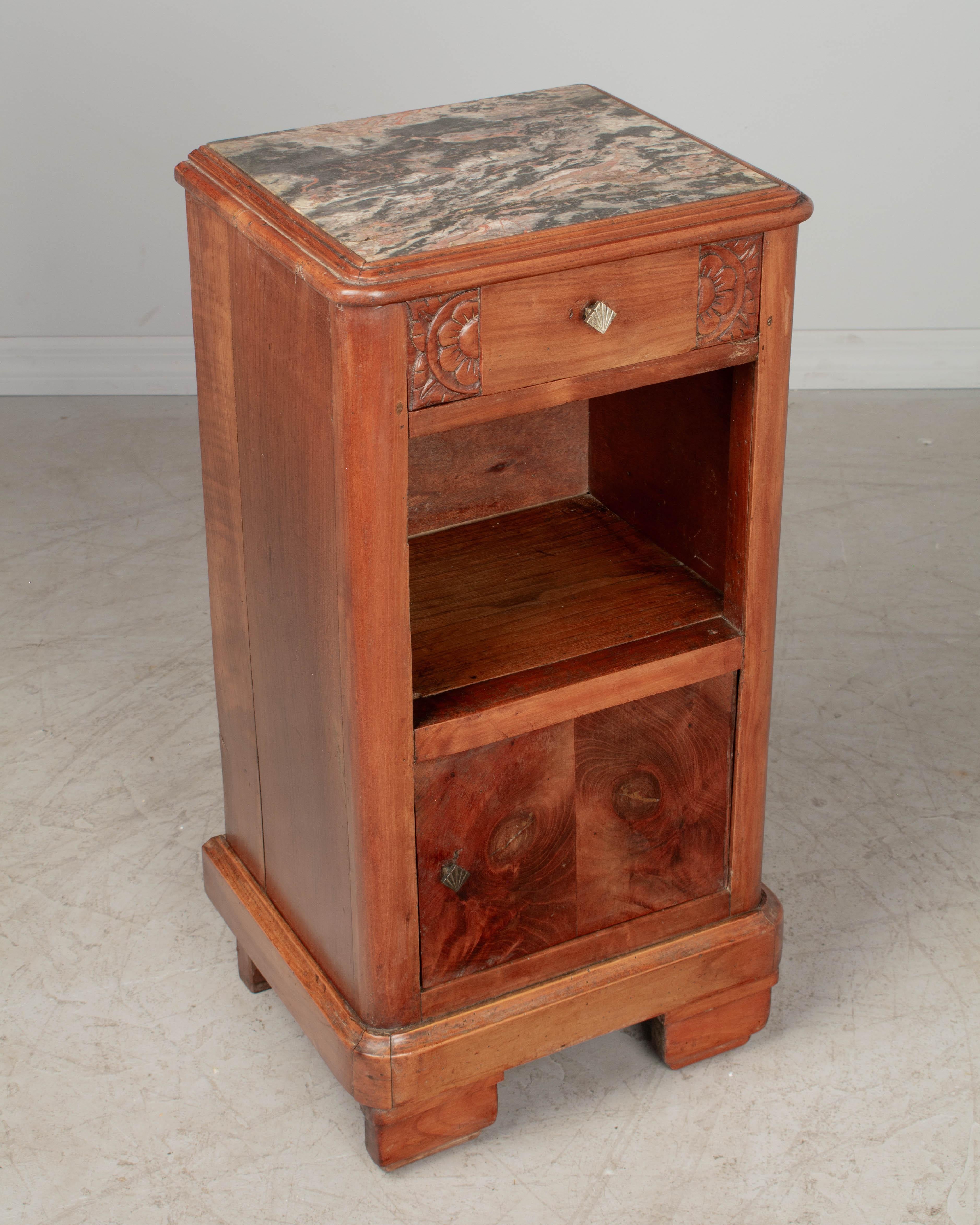 A French Art Deco nightstand made of solid cherry wood, with inset marble top. Dovetailed drawer with hand-carved stylized floral details. Open niche above a cabinet door with bookmatched walnut veneer panels. Nickel plated cast brass knobs. Good