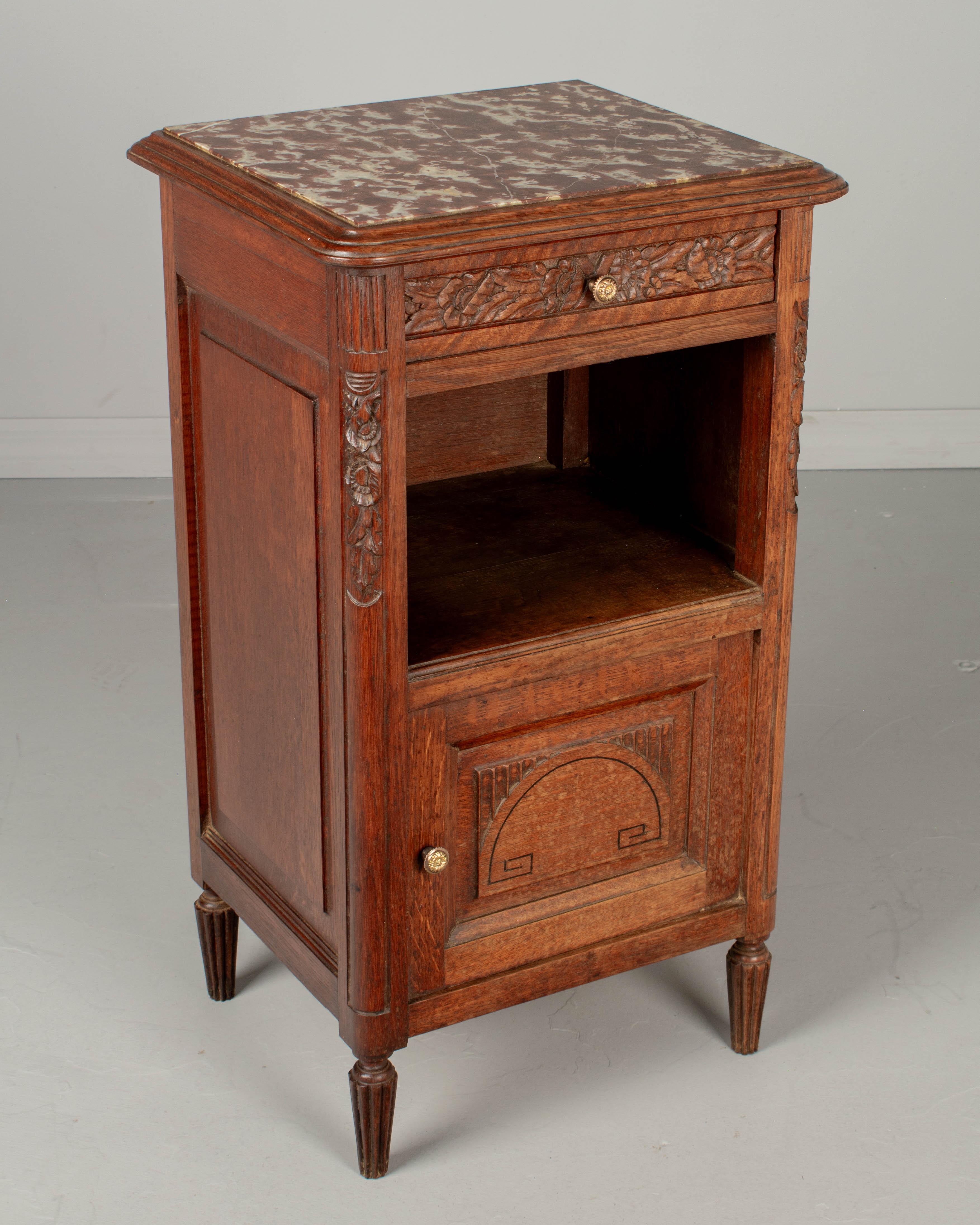 A French Art Deco nightstand made of solid oak, with inset Rouge Royal marble top. Dovetailed drawer with hand-carved stylized floral details. Open niche above a cabinet door opening to a porcelain lined interior. Cast brass knobs. Turned tapered