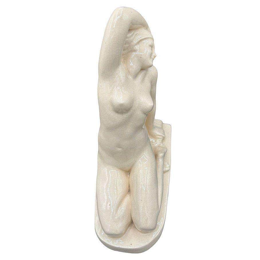 Original Art Deco nude ceramic sculpture featuring a figural study of a young female form with an early streamlined form by F. Trinque
The artwork showcases the woman in pristine white porcelain, positioned on the ground with her thighs together and