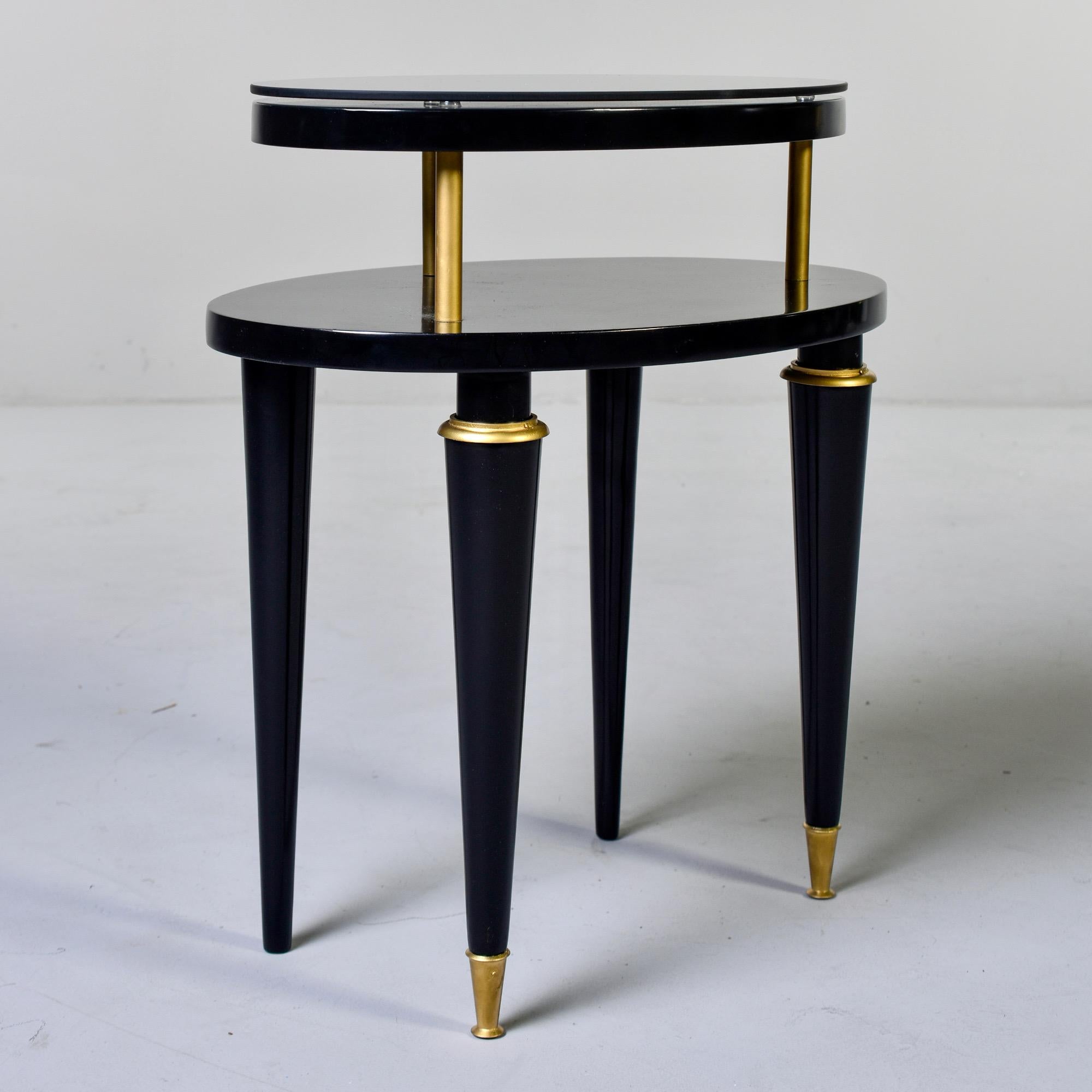 French two tier side table features an oval shaped table top with tapered legs, brass trim and brass supports with a smaller top tier and dark glass oval top, circa 1940s. Unknown maker. New, professionally applied black satin finish.