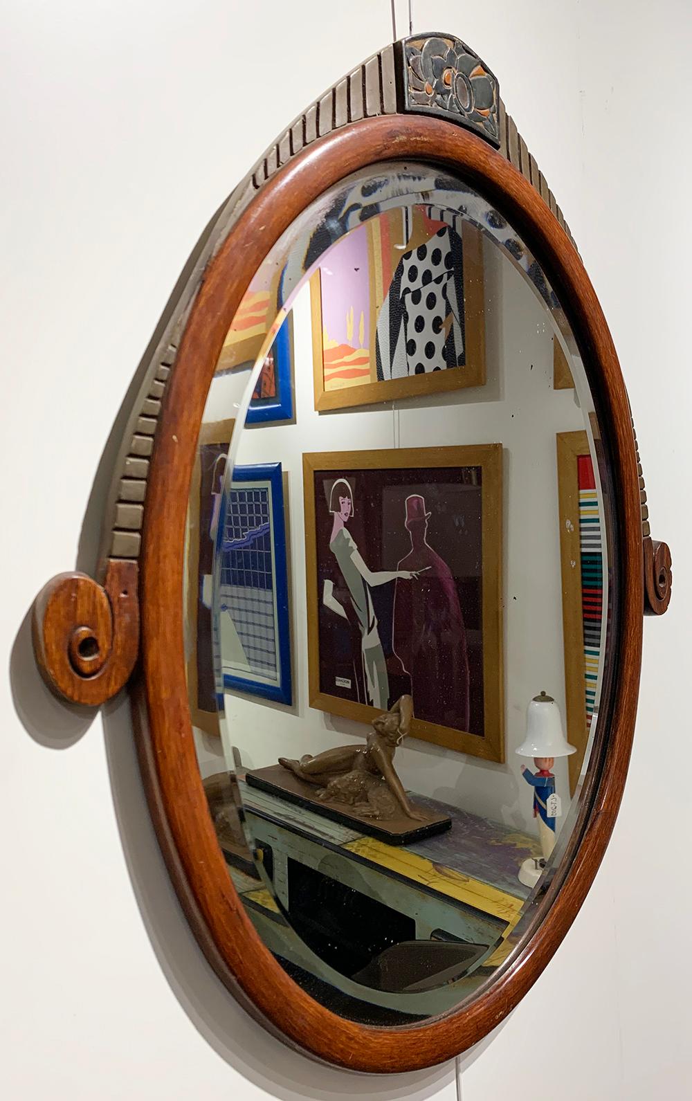 Eleganter französischer Art-Déco-Wandspiegel aus der Mitte des 20. Jahrhunderts. 
Der länglich-ovale Spiegel hat einen abgeschrägten Rand und befindet sich in einem Holzrahmen, der mit stilisierten Blumenmotiven verziert ist. Der Spiegel ist