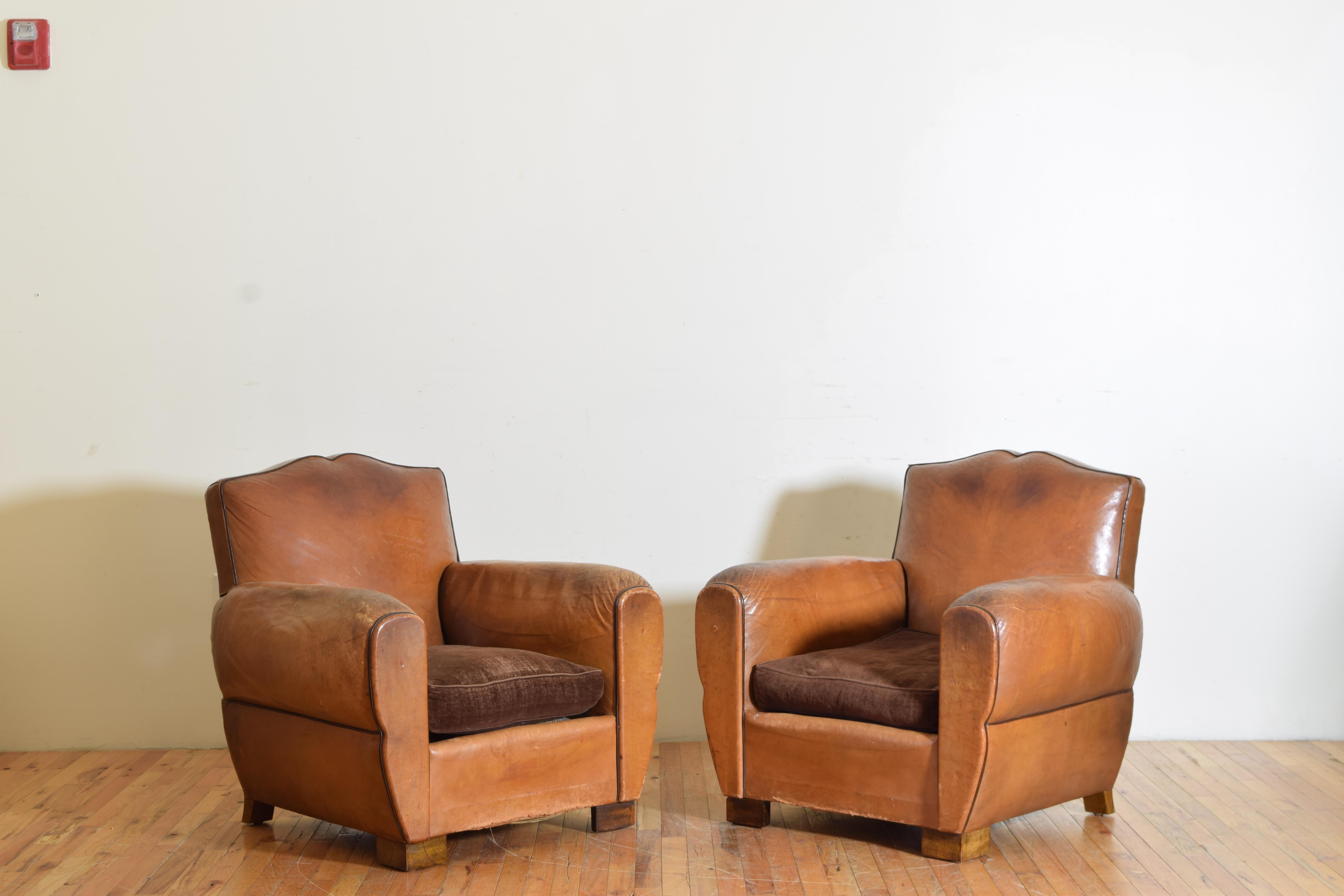 A pair of Art Deco period leather club chairs with black leather piping, brass nailheads, and dark brown velvet down cushion seats. The tops of the chairs are mustache shaped adding to their French style.