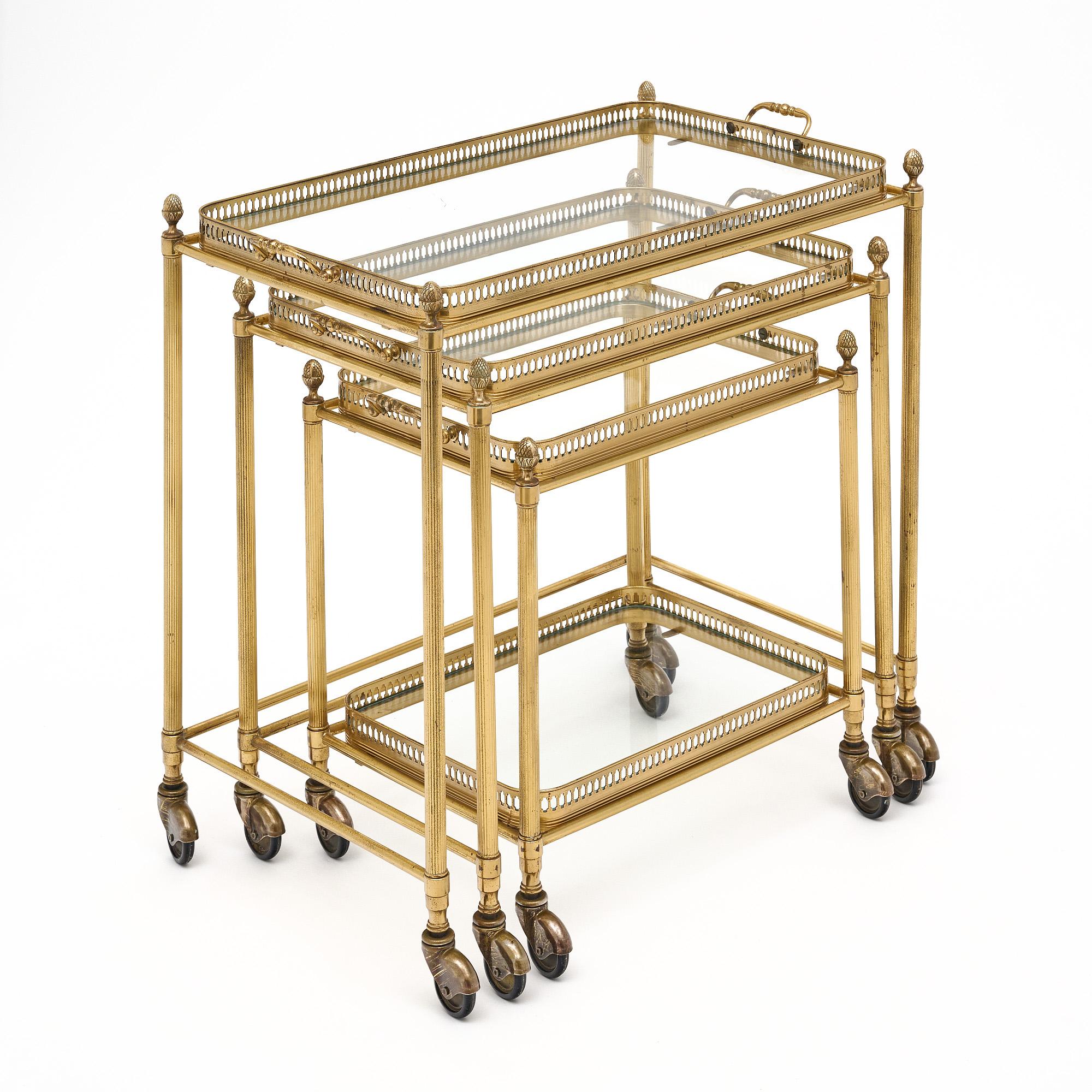 Set of nesting tables or nesting bar carts from France made of finely cast brass. Each of the three tables feature a removable tray with glass bottom, while the smallest table also has a lower glass shelf. Each has stylized acorn finials and