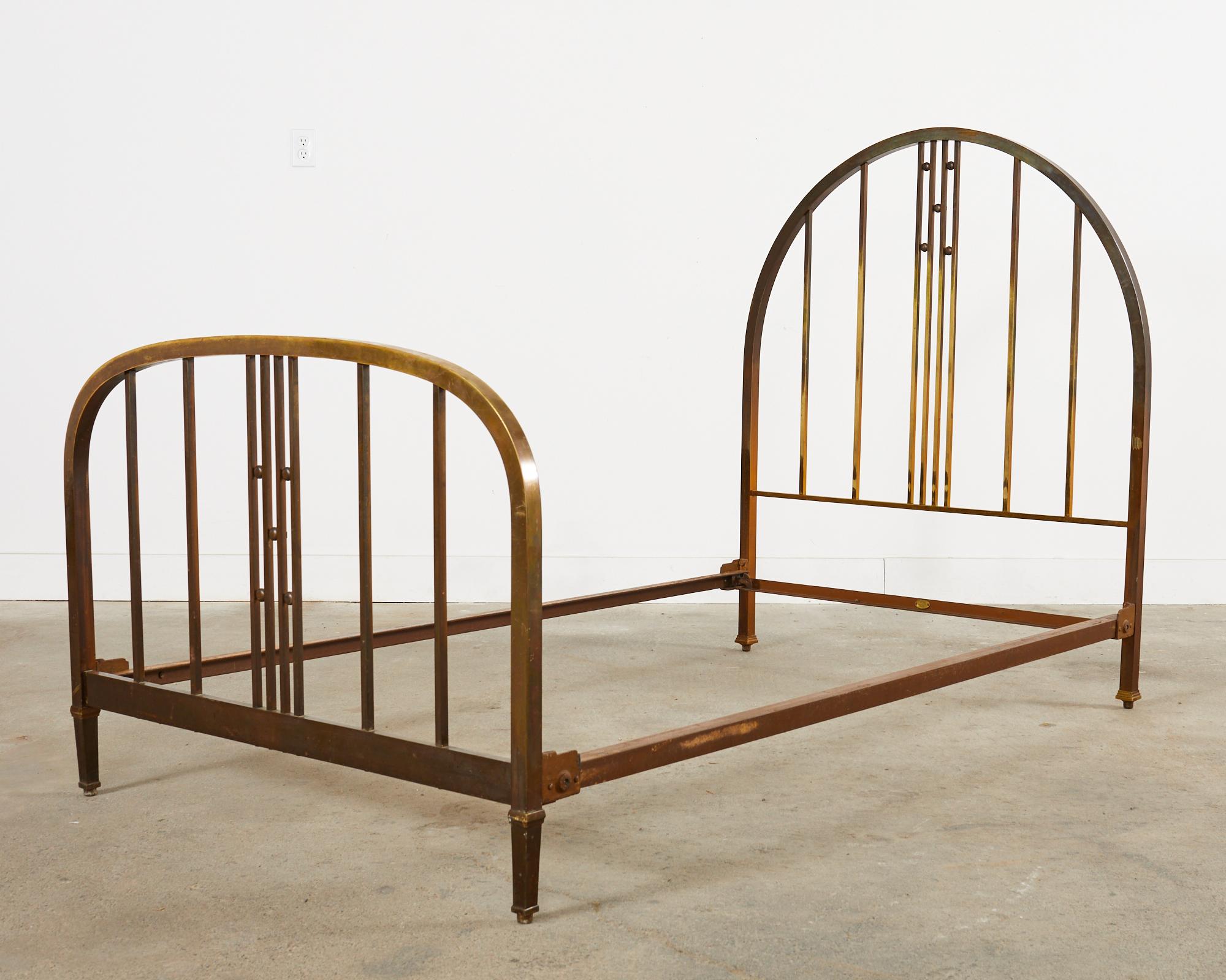 Gracefully curved French art deco bed frame constructed from patinated brass and iron. The bed features a cathedral arch formed headboard having vertical slats decorated with brass orbs or spheres. The curved footboard has a similar design of slats