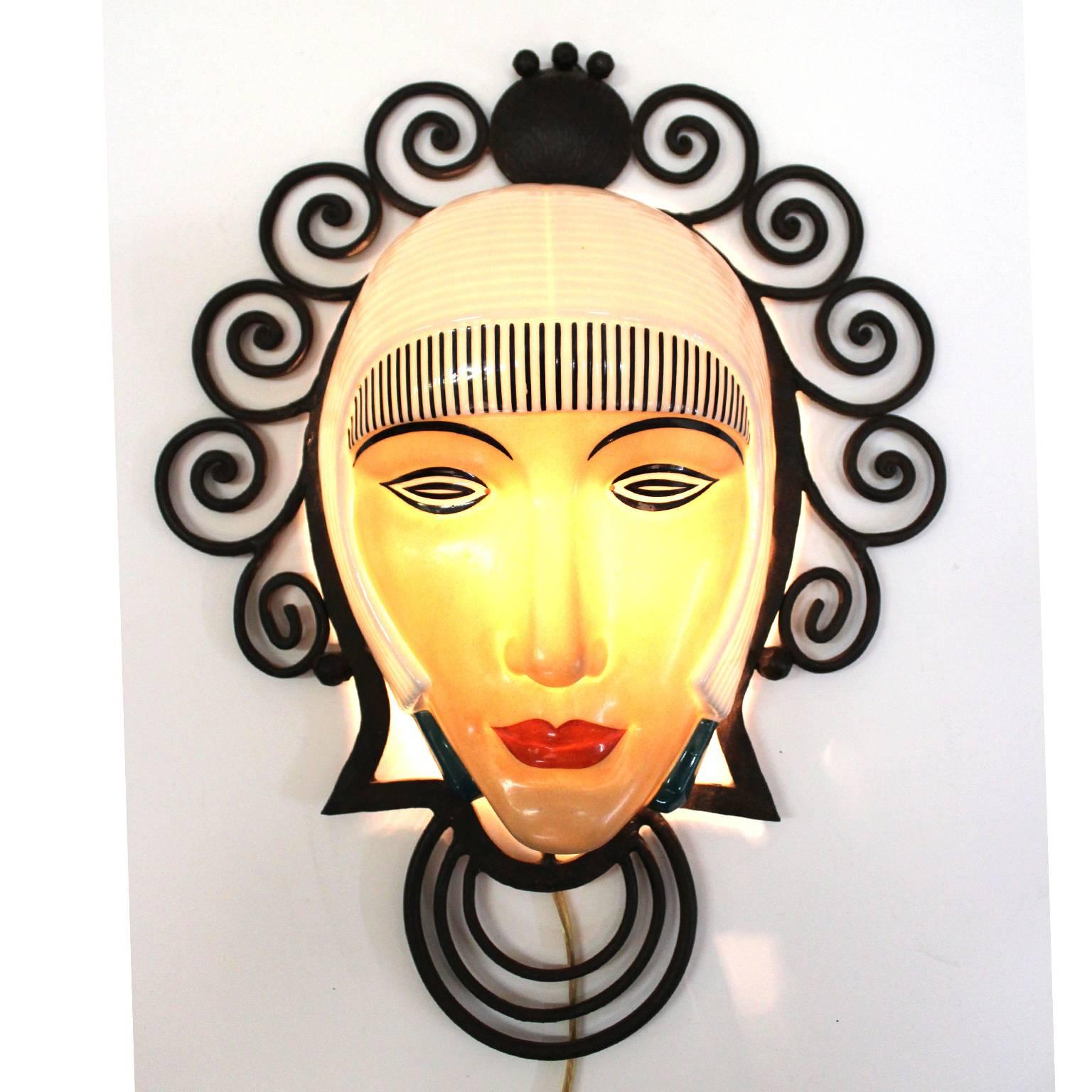A unique 1920s French Art Deco wrought iron sconce with an enameled porcelain mask. The mask face lights up when the light is turned on. No signature, good condition, with slight undetectable hair line on right side.