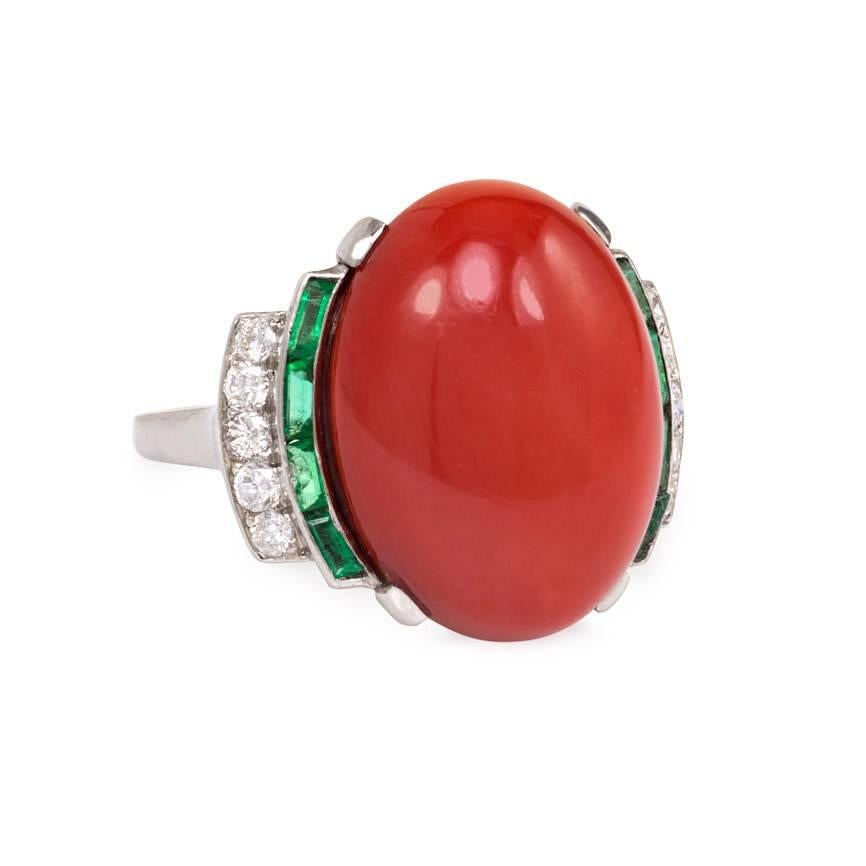 An Art Deco oval cabochon coral ring flanked by emeralds and diamonds, in platinum.  France.  Coral measures approximately 17 x 12.5 x 7.5mm, atw 8.81 ct.