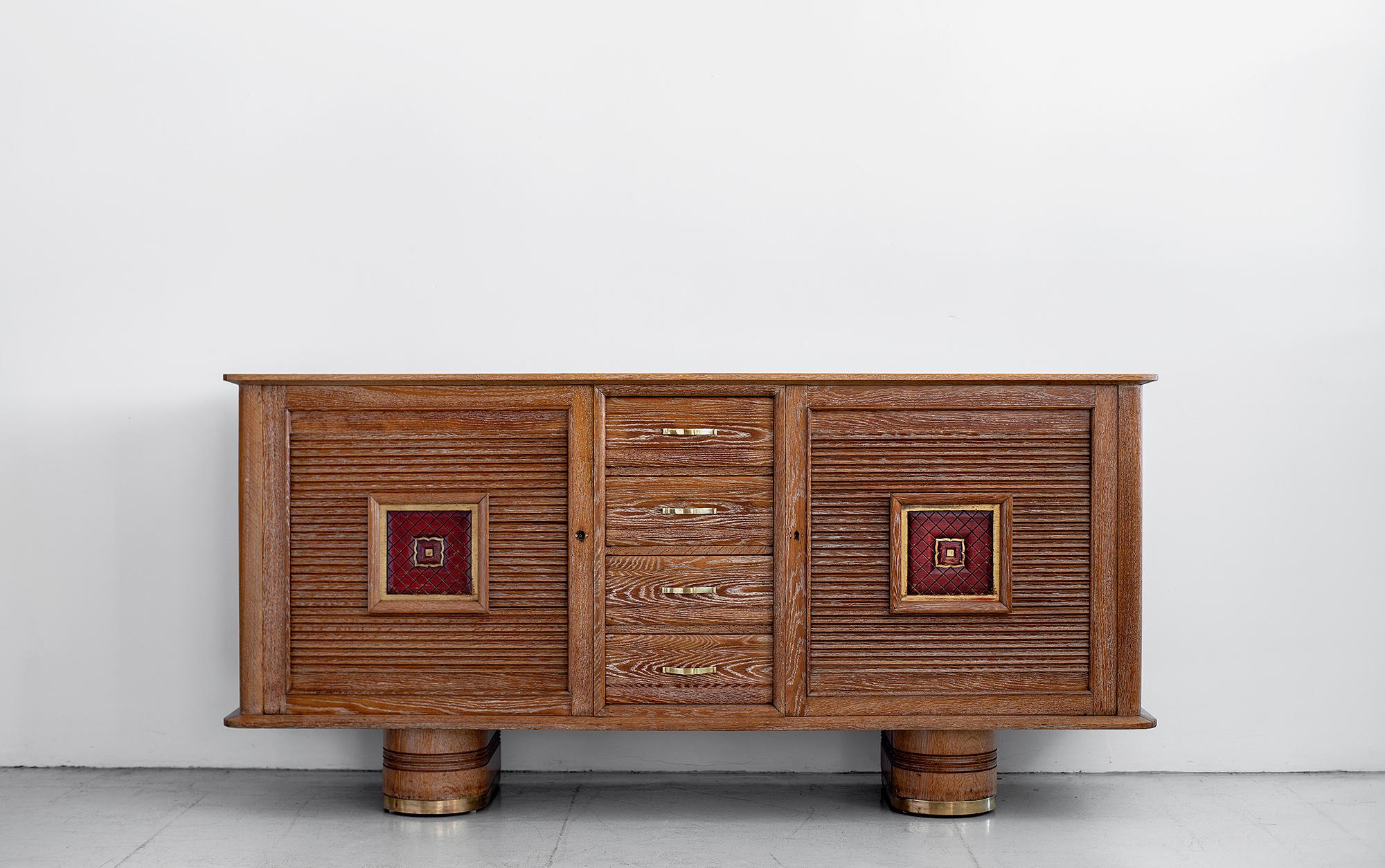 Beautiful Art Deco 1940s French cerused oak sideboard. 
Sits on 2 pontoon legs with brass four drawers flanked by two doors with open storage. 
Undulated wood detail with red leather. 

   