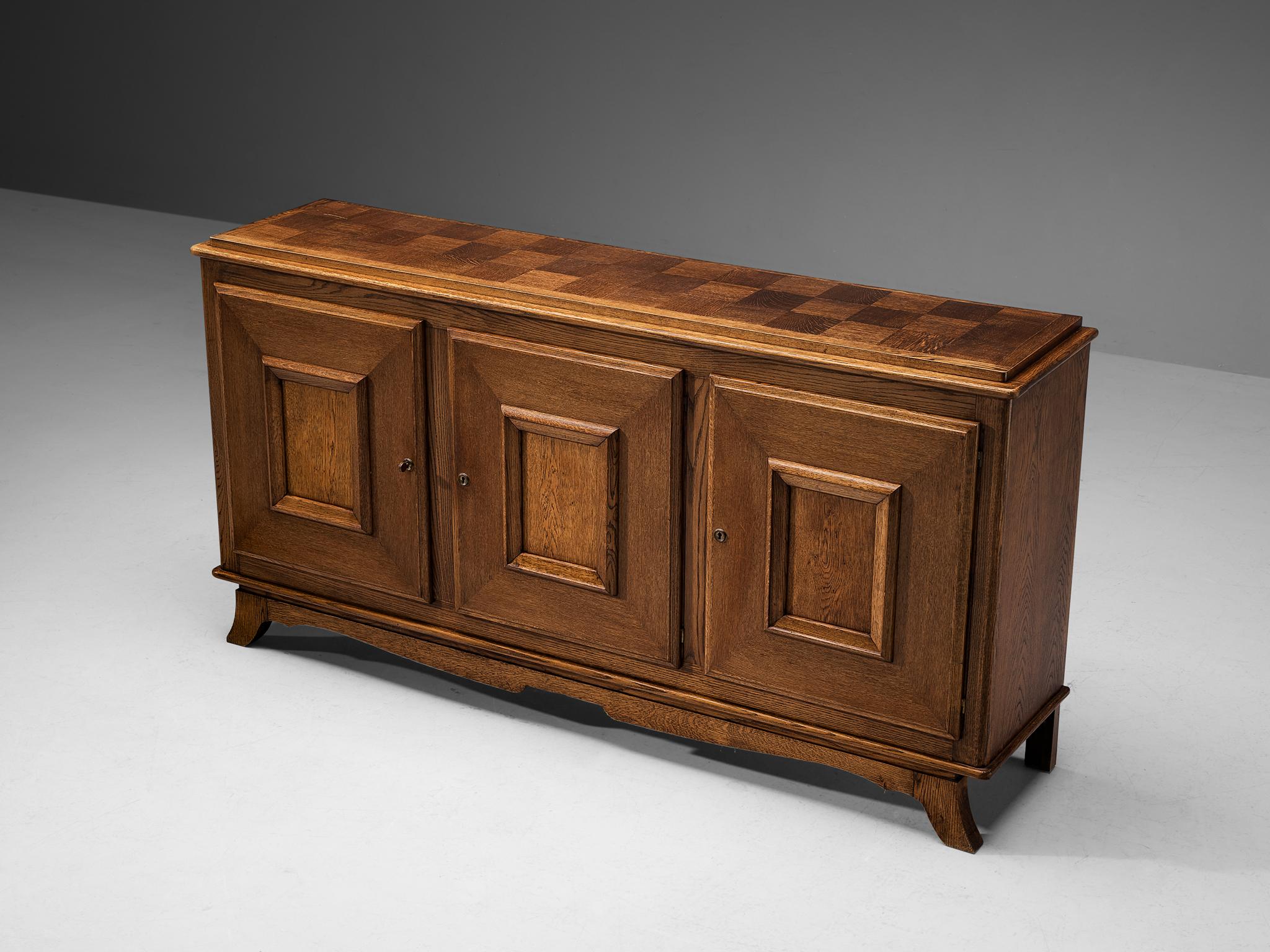 Sideboard, oak, France, 1940s

This sideboard made in oak has very nice and distinct features. For example the inlayed checkered top, the rectangular panelling on the three doors, and the tapered front legs of the piece that point outwards. Also,