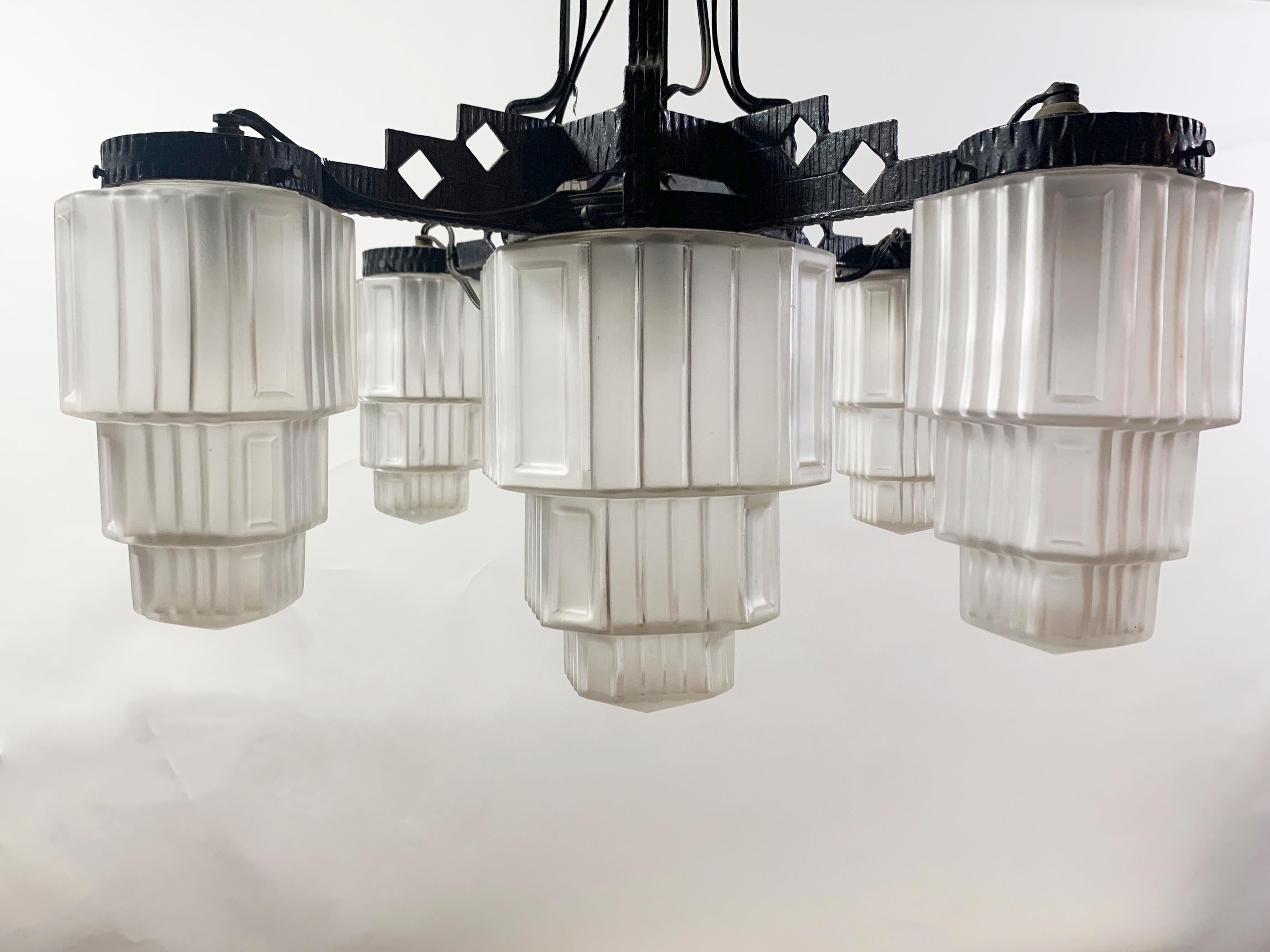 Questo squisito lampadario cattura l'essenza della raffinatezza dell'Art Déco degli anni '20-'30. Realizzato con una struttura in ferro battuto forgiato, presenta audaci motivi geometrici emblematici del periodo. Il lampadario presenta cinque