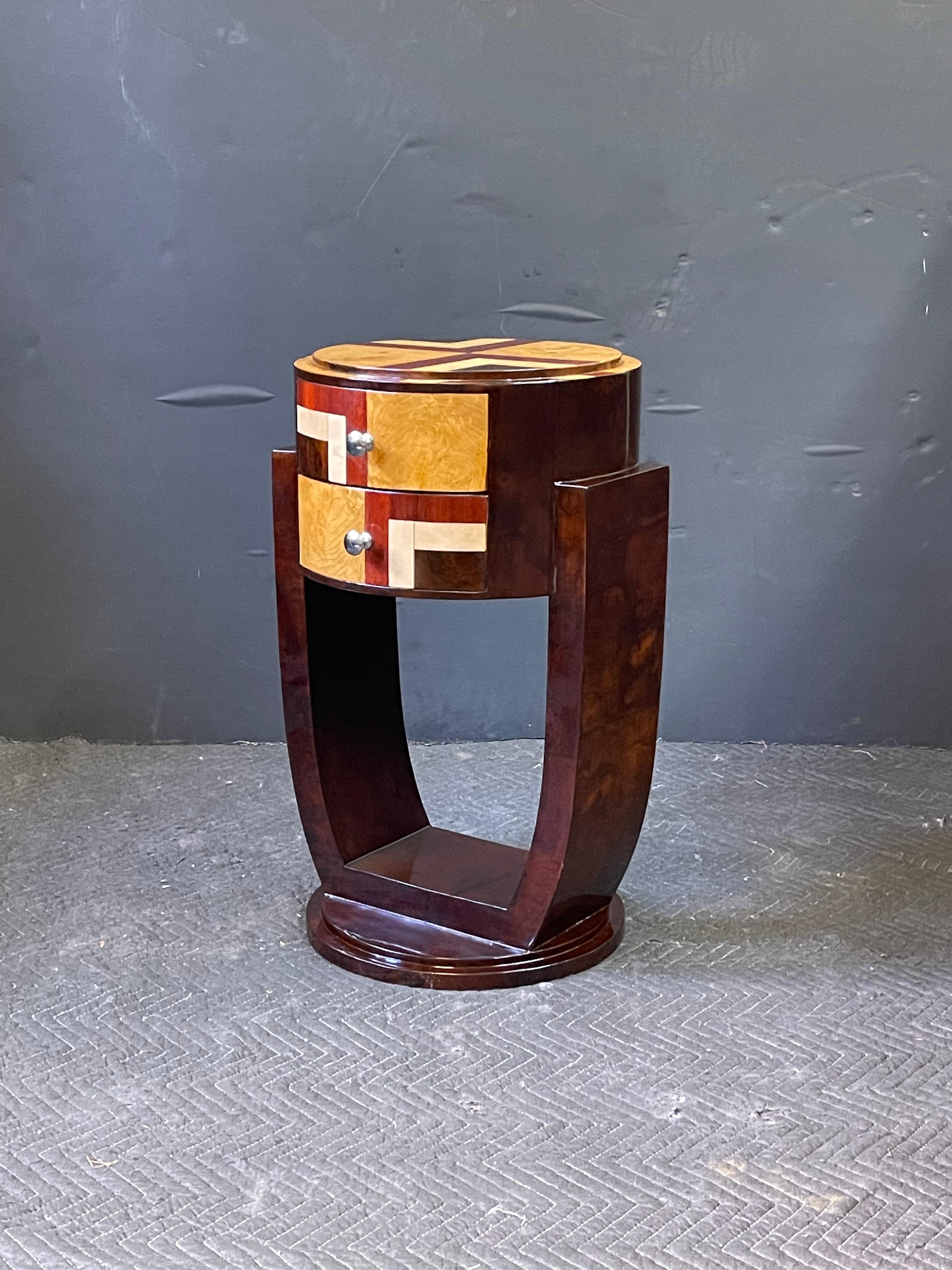 This mid 20th Century French side table is in the streamlined style of Art Deco and features prominent inlaid veneers of jacaranda, maple, and walnut. The circular table has a top of geometric inlay and steps down to a frieze containing two drawers