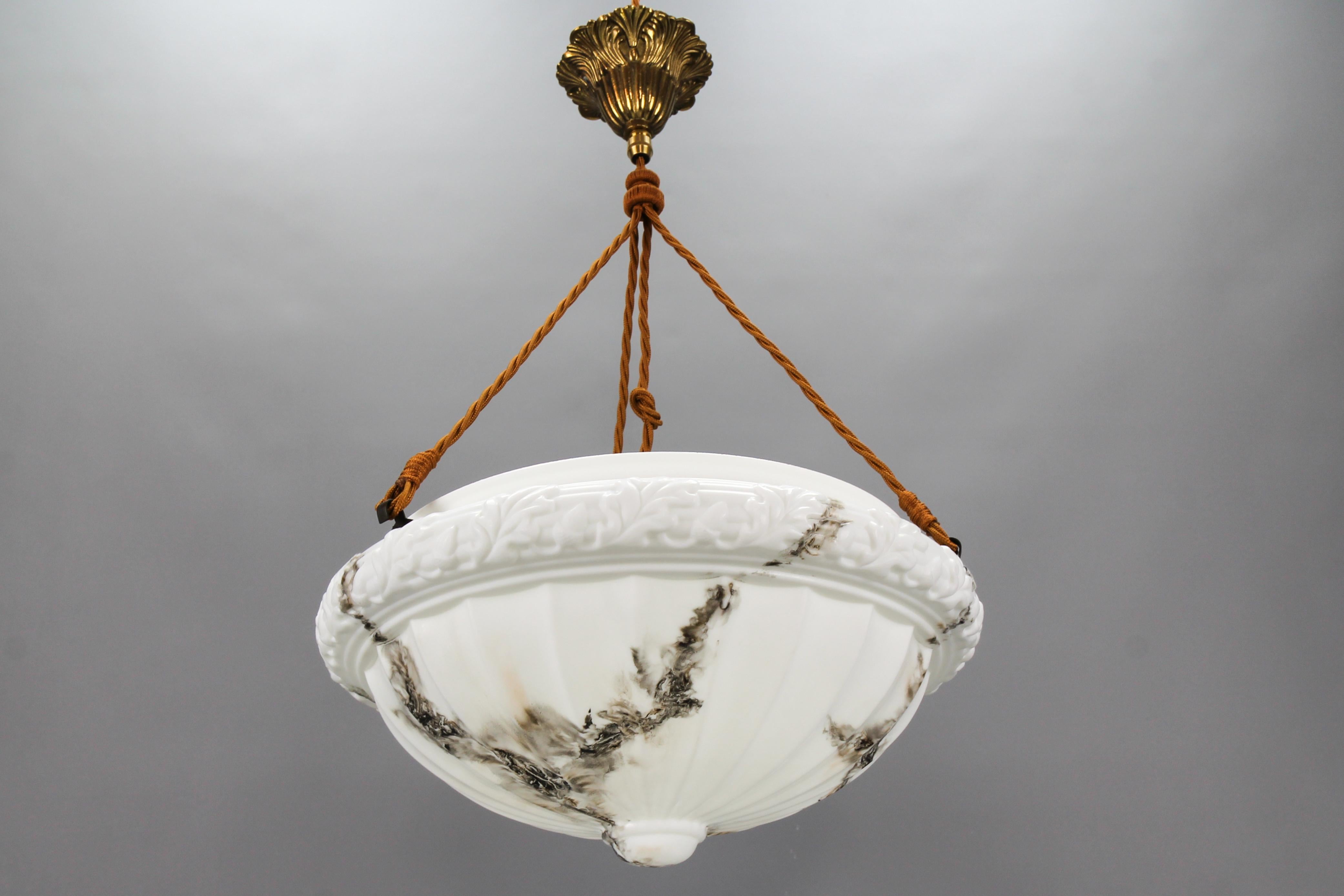 An Art Deco style marbled milk glass and brass pendant light fixture from circa the 1950s, France.
This beautifully shaped ceiling light fixture features an alabaster effect milk glass lampshade with a relief design – a pattern of acorns and oak