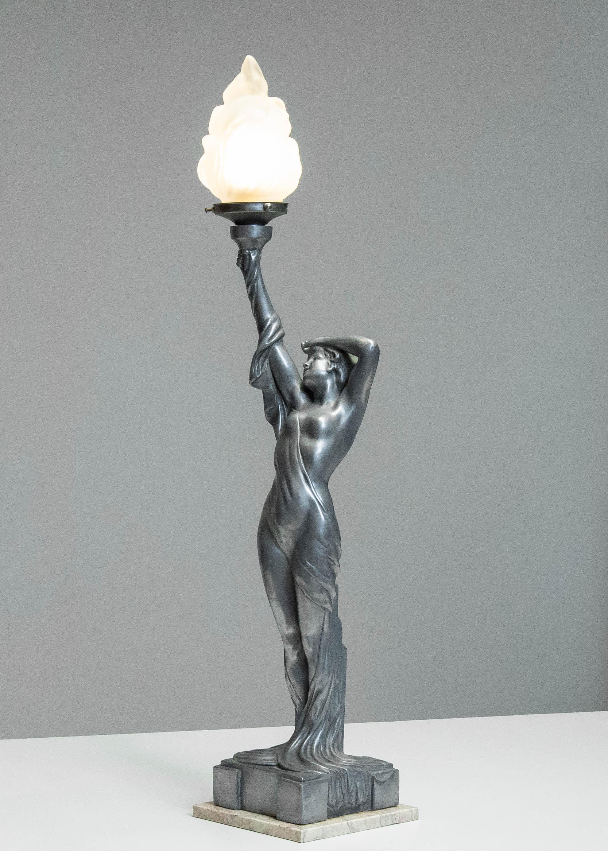 Lampada da tavolo in stile Art Déco con una figura femminile scultorea in un fluente abito drappeggiato, che regge una torcia con un paralume a forma di fiamma in vetro smerigliato. Realizzato in lega di zinco-alluminio con finitura patinata,