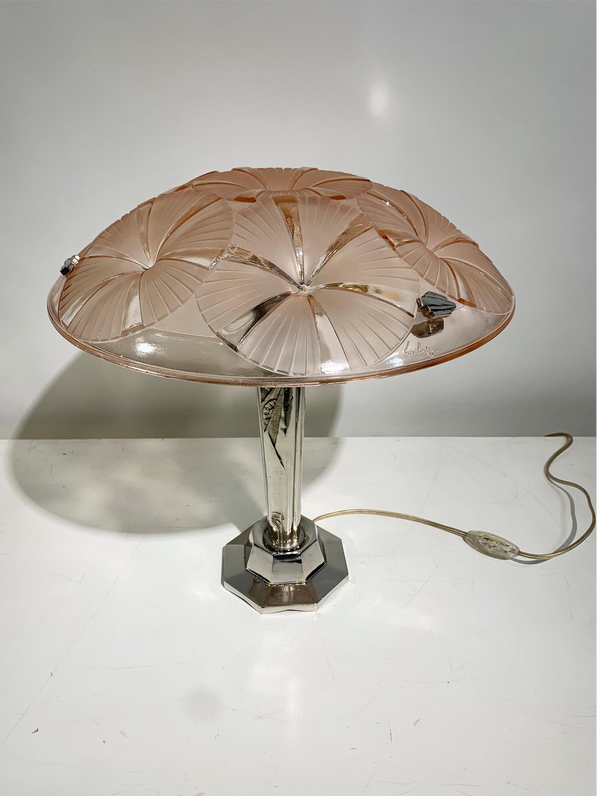 Esta exquisita lámpara de mesa Art Déco, firmada  Degué, presenta una hermosa pantalla de cristal moldeado con intrincados motivos geométricos y florales estilizados en un suave tono rosa, característico del diseño francés de los años 1920-1930. 
La