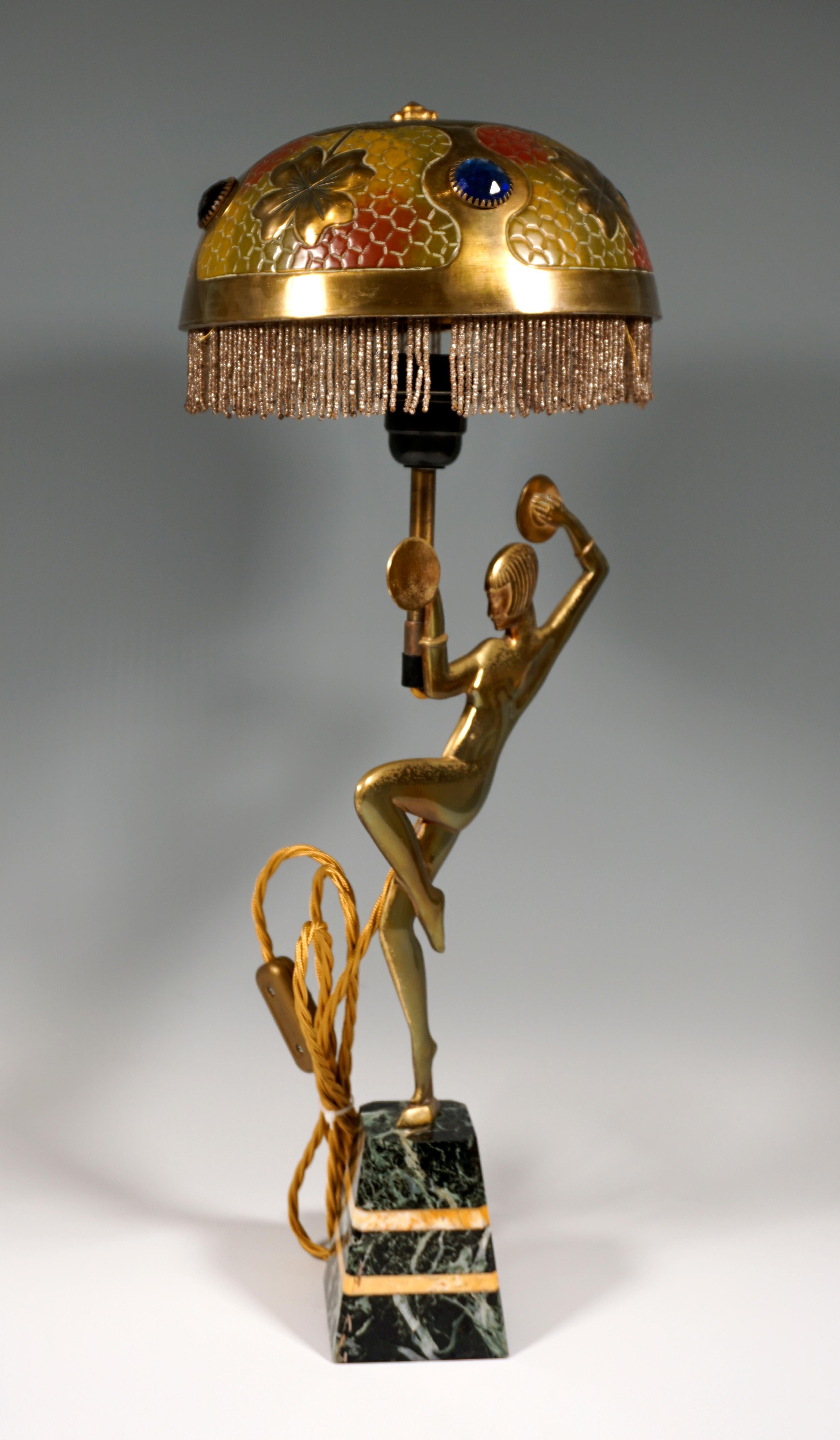 Table lamp with a flat, relief-like, unclothed dancer standing on one leg with cymbals on a black and yellow marble base in the shape of a truncated pyramid. The dome-shaped lampshade with pearl fringes gives a floating impression due to the