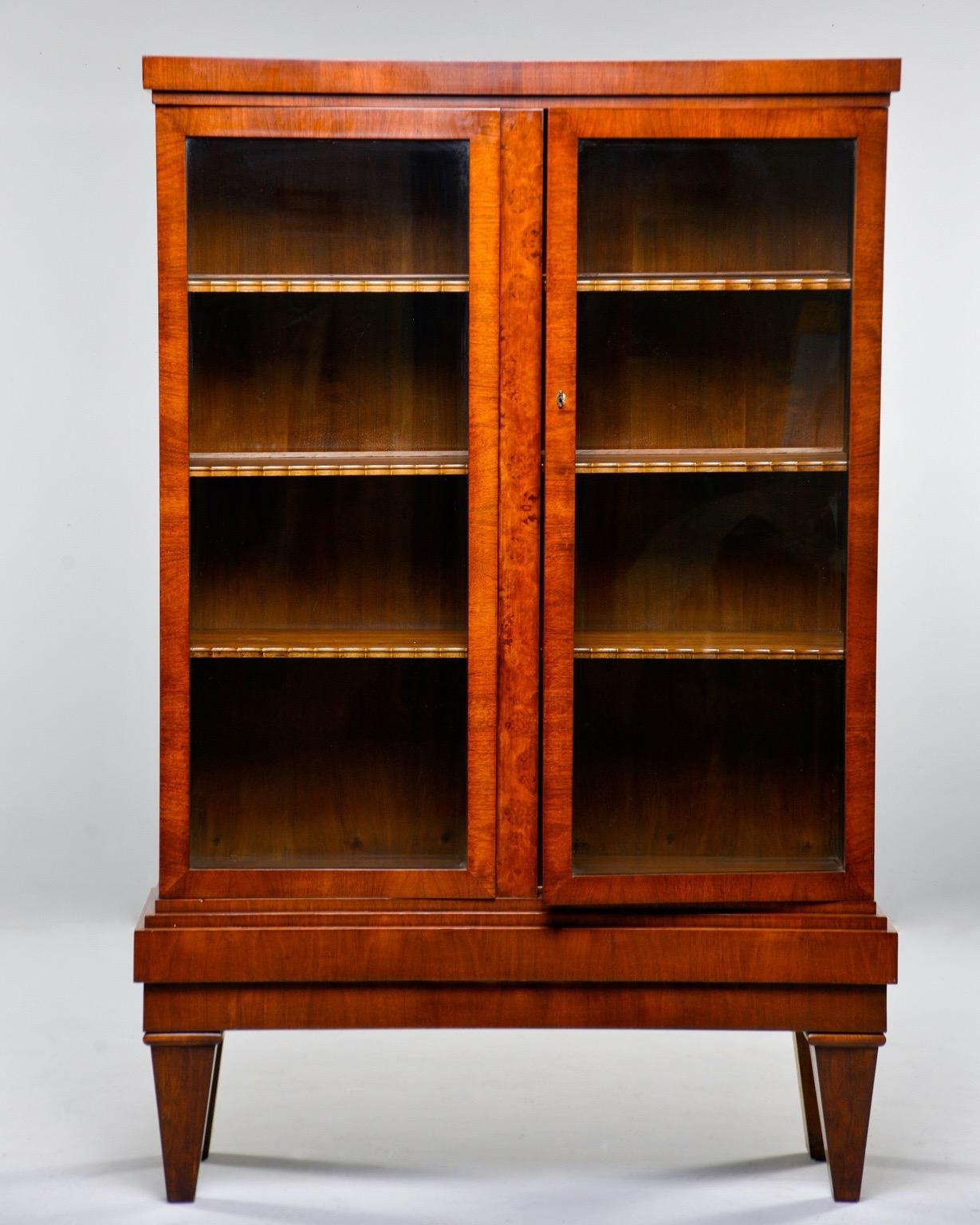 French cabinet or bookcase is nearly 6 feet tall and features two glass-fronted hinged doors that open to compartment with three fixed shelves and sturdy, stepped base, circa 1930s. Unknown maker.