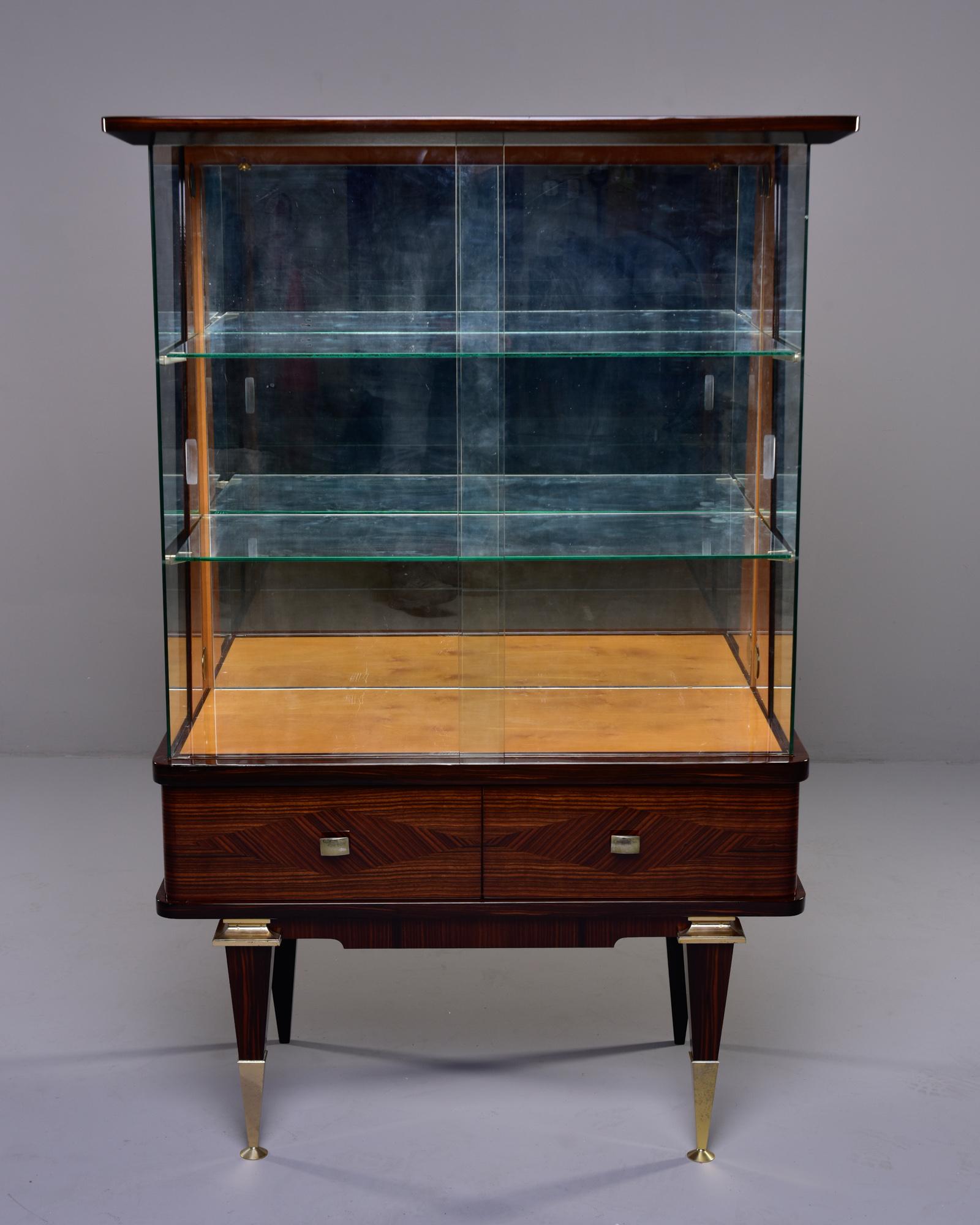 Circa 1940s French Art Deco vitrine in macassar ebony with marquetry pattern and contrasting maple interior trim. Brass trimmed legs. Sliding glass doors with two adjustable interior glass shelves and mirrored back. 

Very good vintage condition