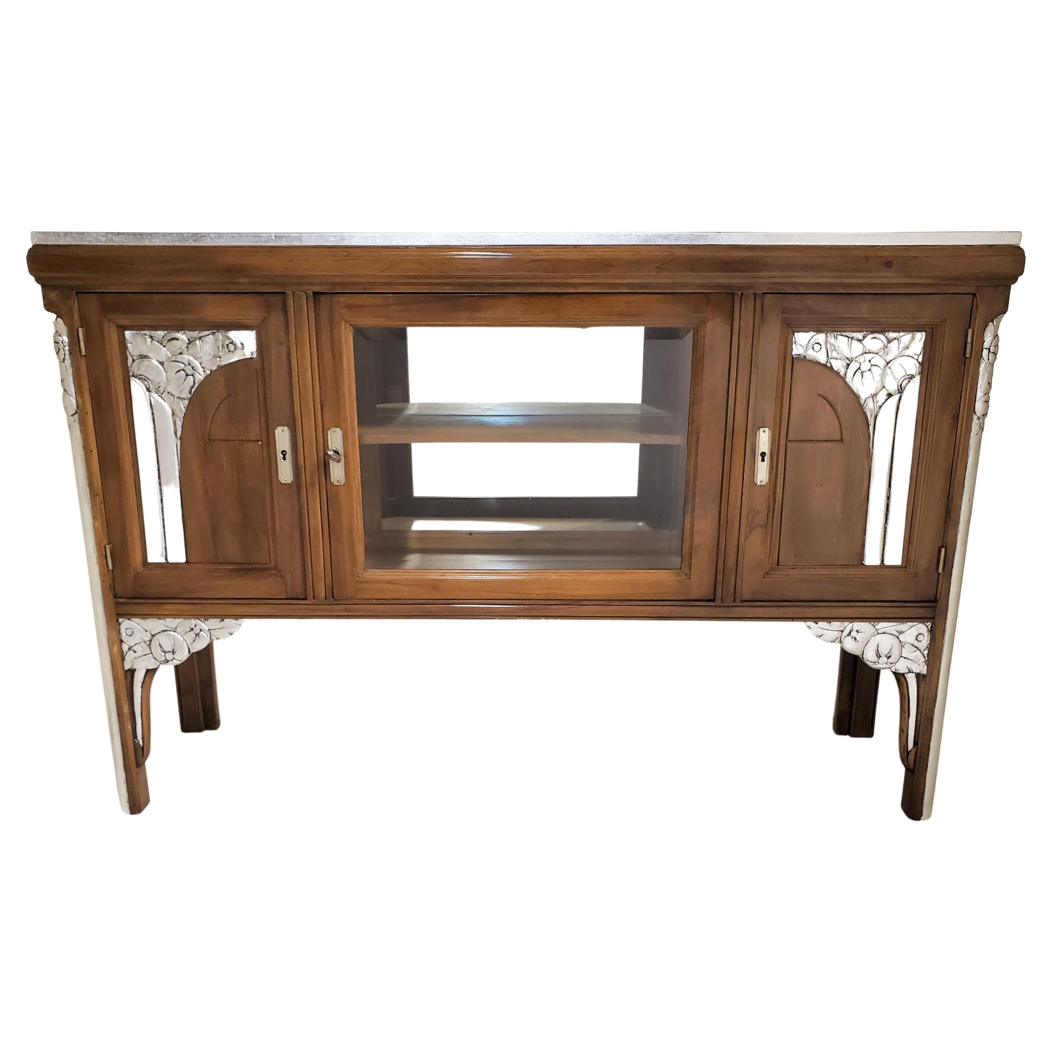 A French Art Deco three door walnut cabinet with hand carved metal leaf detail of geometric and stylized floral design. The walnut wood in a rich, mellow color adds warmth to the piece.
The center of the cabinet houses a mirrored backed glass