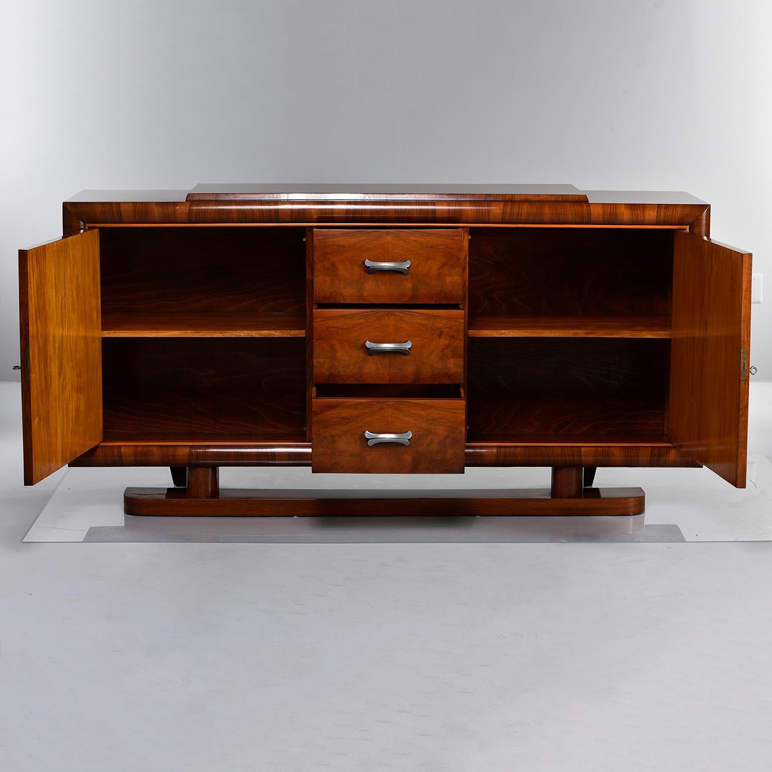 French deco era sideboard in walnut with three centre drawers flanked by locking cabinets with single internal shelf, circa 1930s. Details include a pedestal base at front, nickel hardware and bookmarked veneer panels on cabinet doors and cabinet