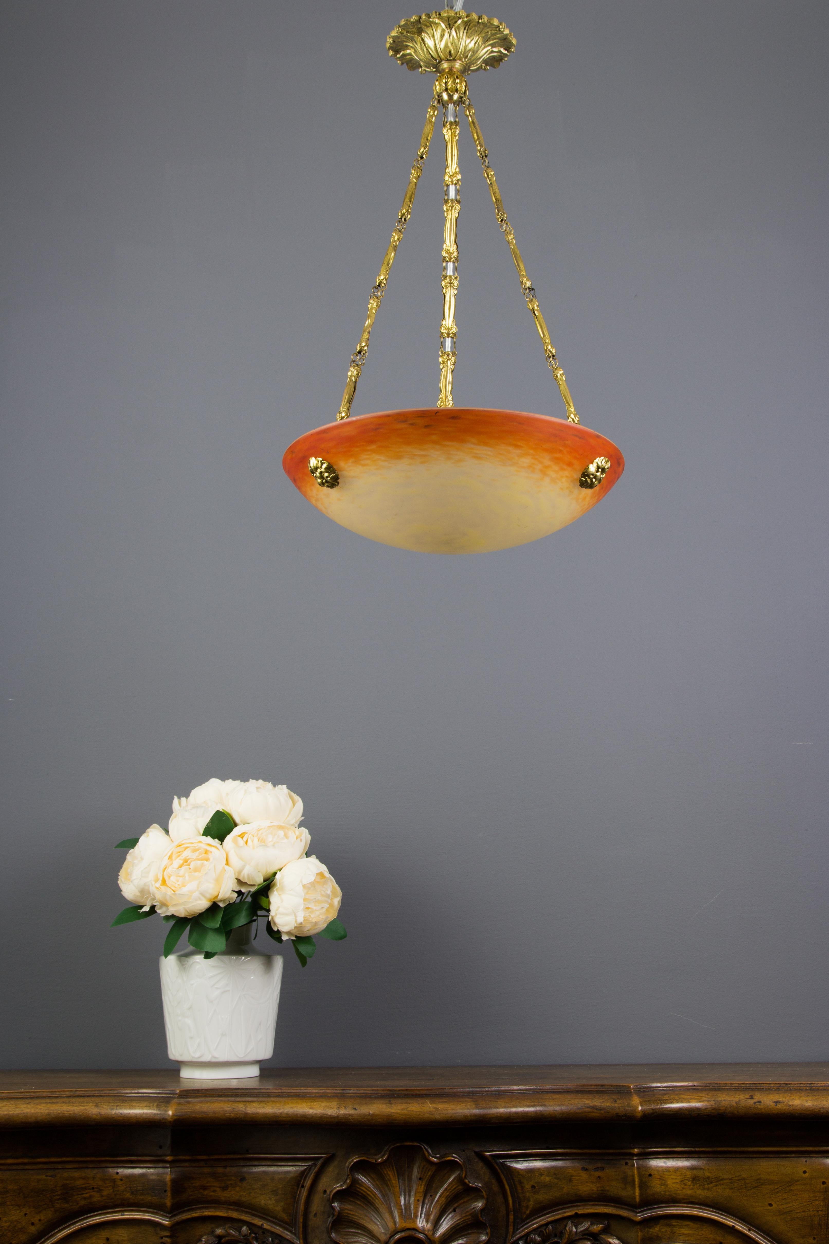 This adorable French Art Nouveau period pendant chandelier features a mottled “Pâte de Verre” art glass shade in orange, yellow, and cream color, signed ”Schneider” by Charles Schneider, hung at an ornate brass and bronze fixture with one socket for