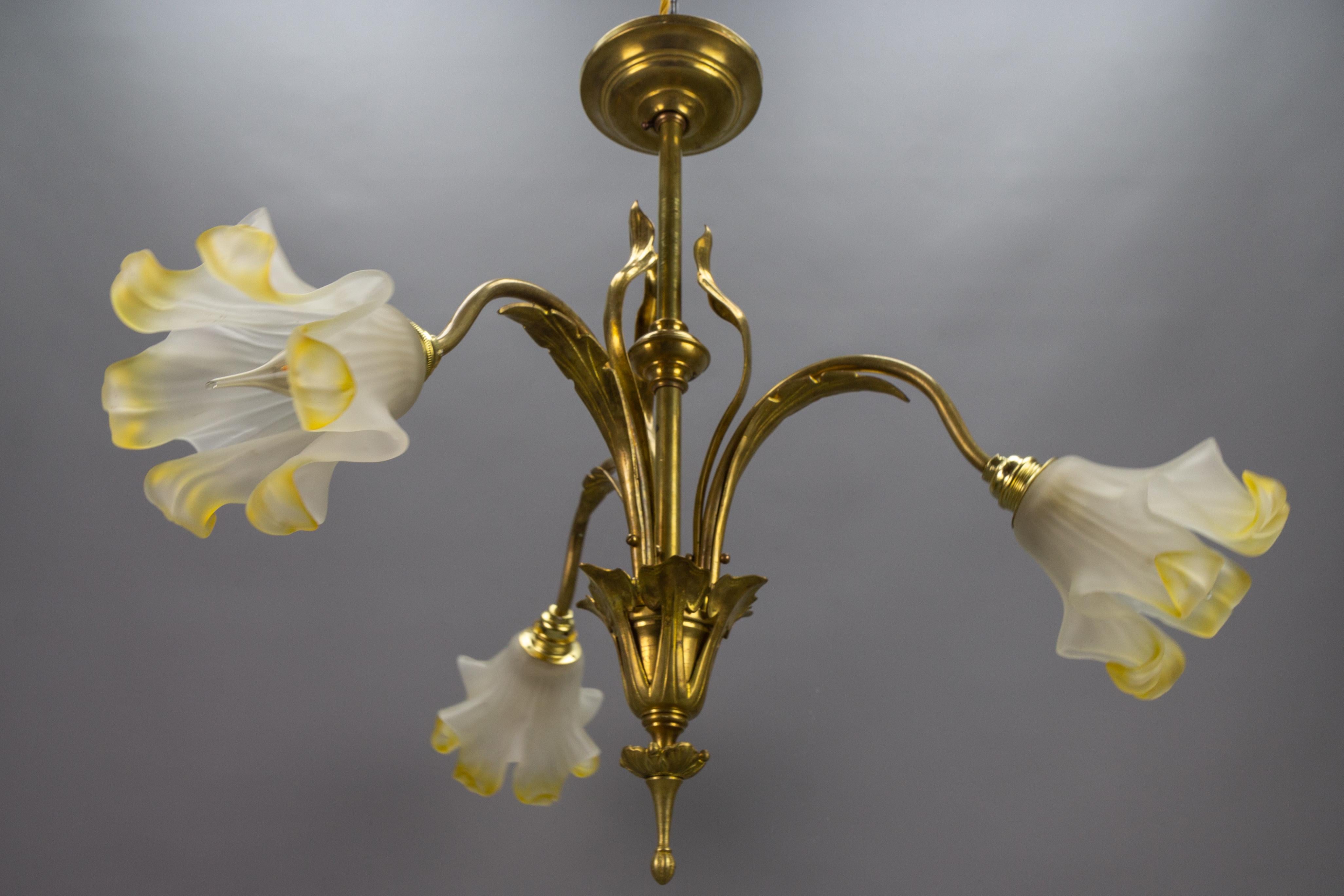 French Art Nouveau brass and glass three-light iris-shaped chandelier, from ca. 1910s.
This absolutely adorable Art Nouveau period brass chandelier features three white and light yellow colored iris-flower-shaped Pâte de Verre glass lampshades and