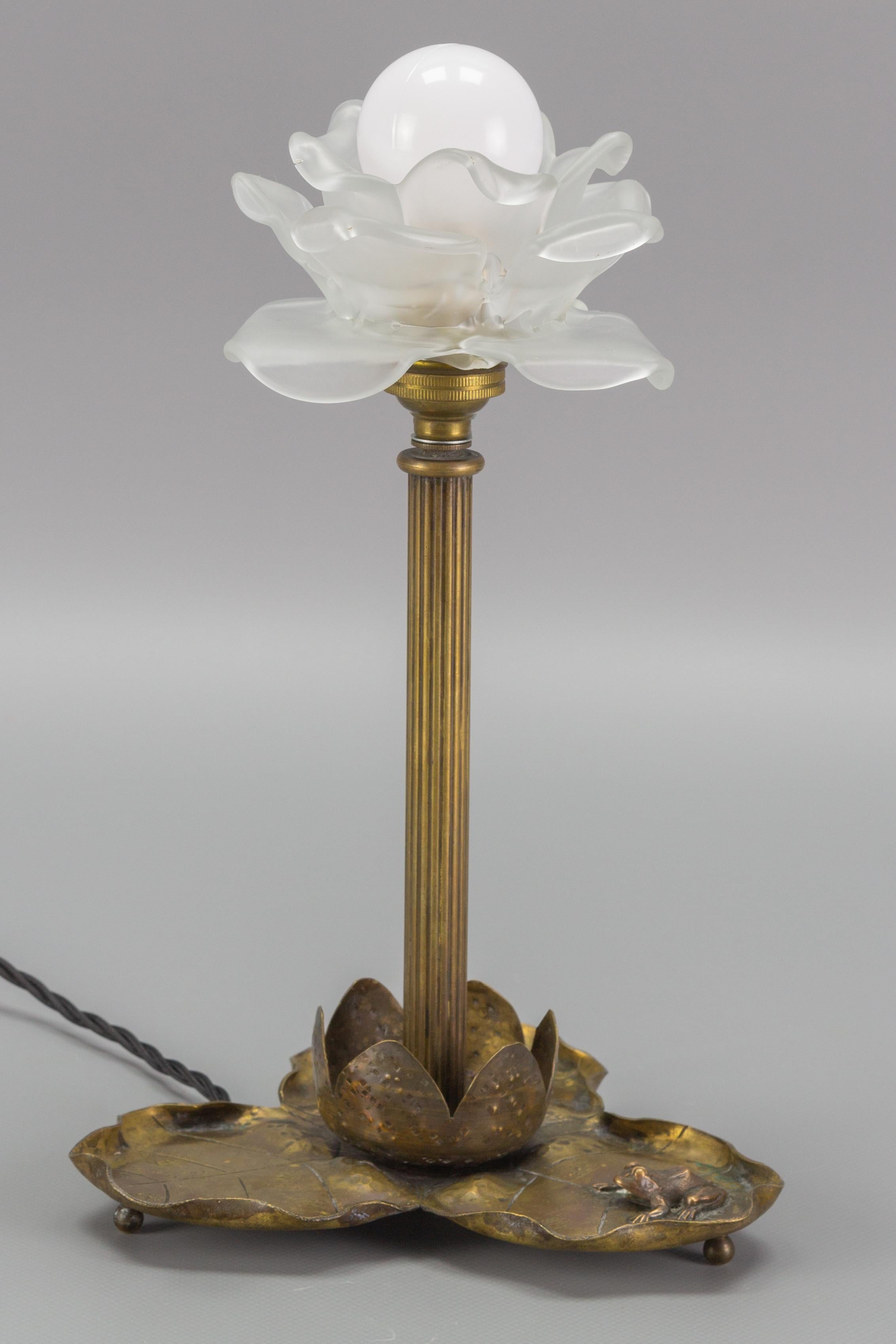 Schöne Jugendstil-Tischlampe aus Messing in Form einer Seerose mit einer Froschfigur auf dem Sockel. Weißer blumenförmiger Lampenschirm aus Milchglas.
Eine Fassung für eine Glühbirne der Größe B22. 
In die USA wird die Lampe mit einem Adapter für
