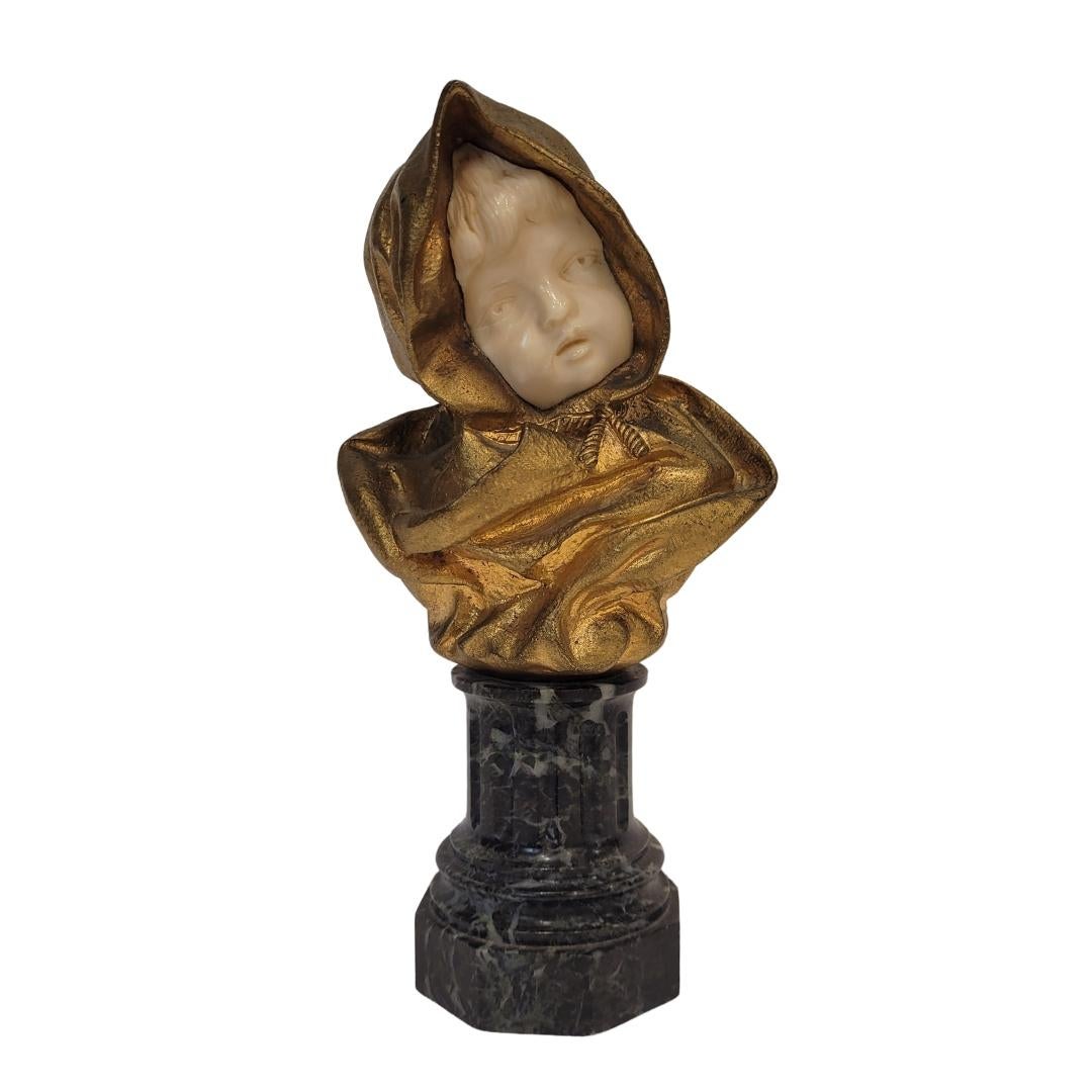 Exquise et très raffinée sculpture chrysoéléphantine représentant le buste d'un enfant enveloppé dans un manteau doré à capuchon. L'œuvre combine l'ivoire sculpté pour le visage, qui émerge de la draperie dorée, créant un beau contraste entre la