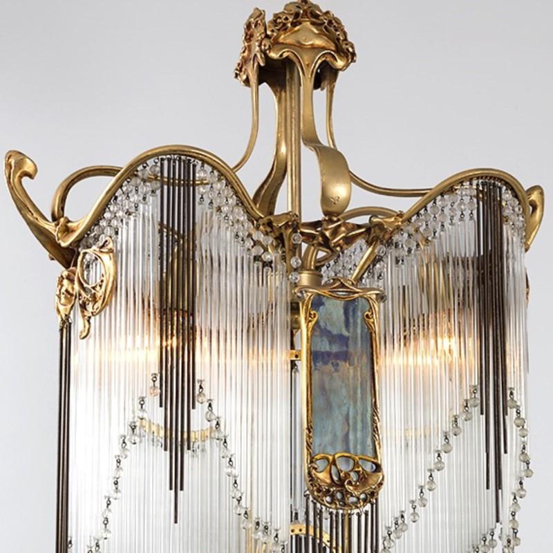 A French Art Nouveau chandelier by Hector Guimard. Dozens of clear glass prisms, each ending in a clear glass bead, hang from a gilt bronze frame with foliate motif. In several places the prisms are interrupted by grouped bronze rods. On each long