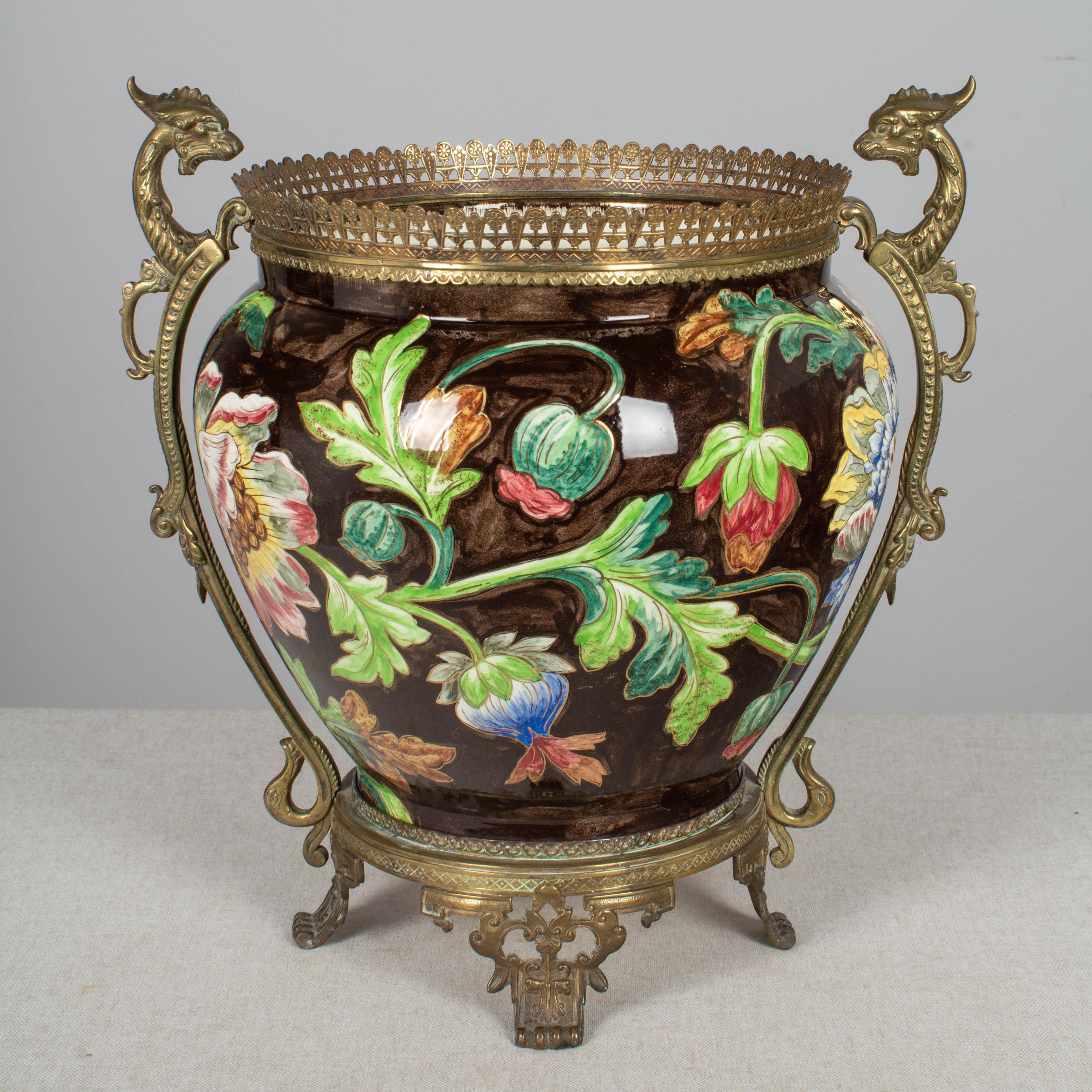 A large French Art Nouveau bronze mounted glazed ceramic planter or cachepot from Longchamp, Burgundy. Vibrant hand painted colorful flowers with bright green foliage and metallic gold accents are dramatically offset on a dark brown ground.
