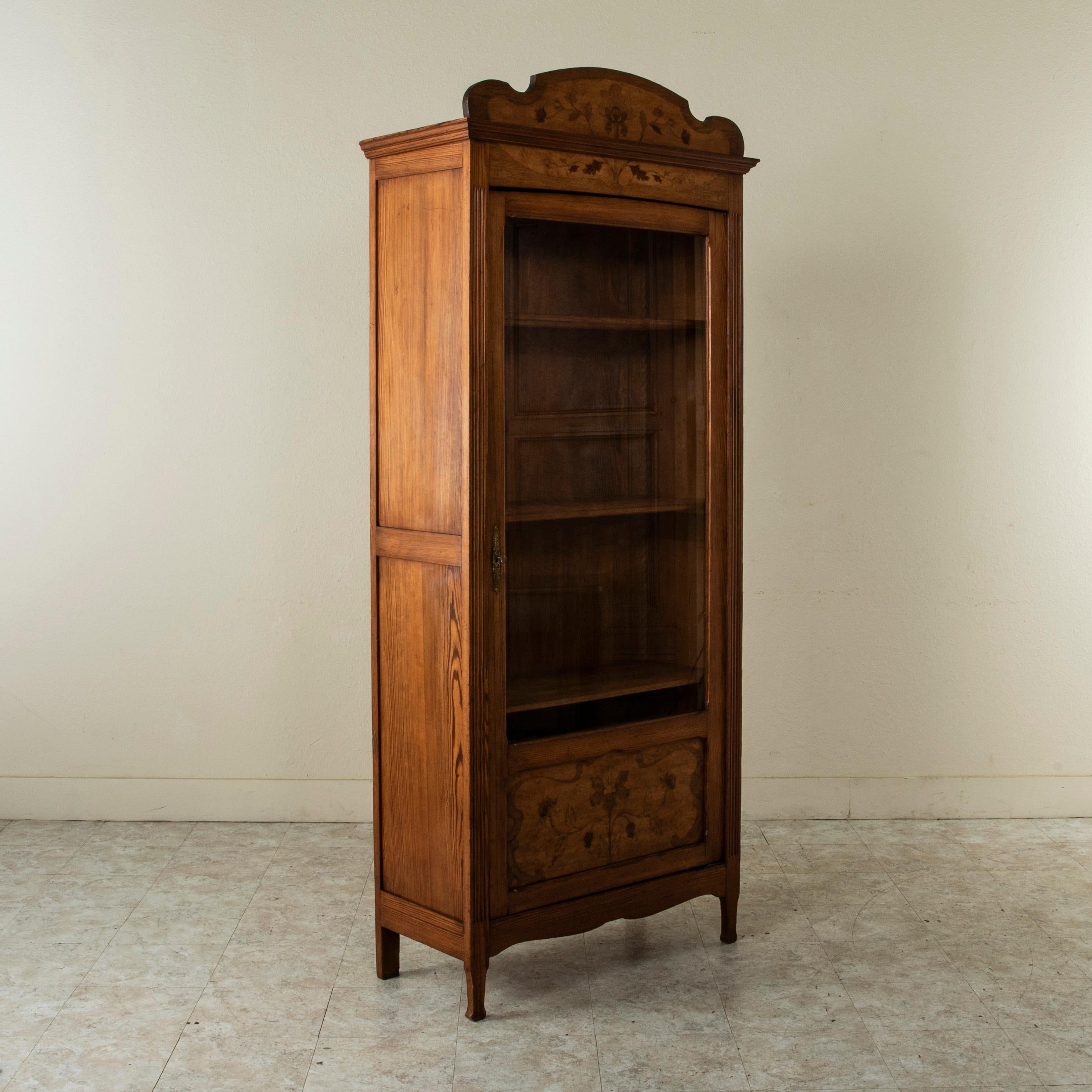 This French Art Nouveau period bookcase or vitrine from the turn of the twentieth century is constructed of solid pitch pine and features a symmetrical inlaid floral motif on the pediment and lower door panel. Fluted columns flank its single door.