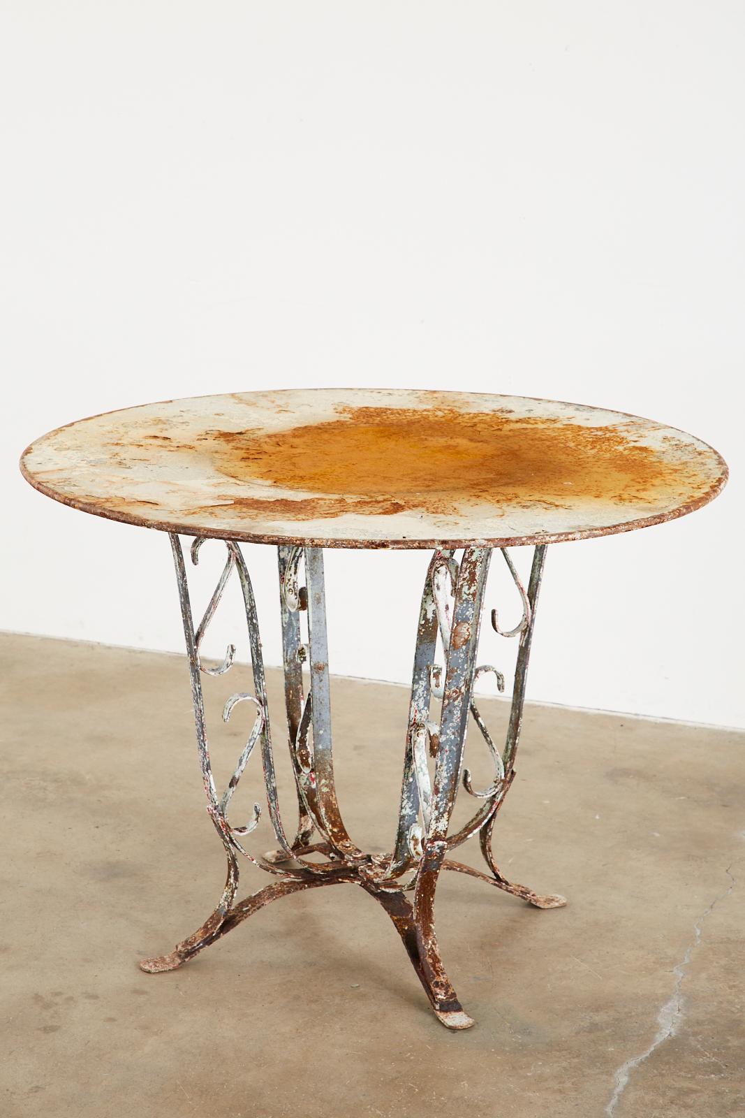 Patinated French art nouveau round garden dining table featuring a sculptural tulip iron base. The round top has rolled edges and is supported by four graceful legs with scroll designs in set. The legs conjoin on bottom and end with flat feet. The