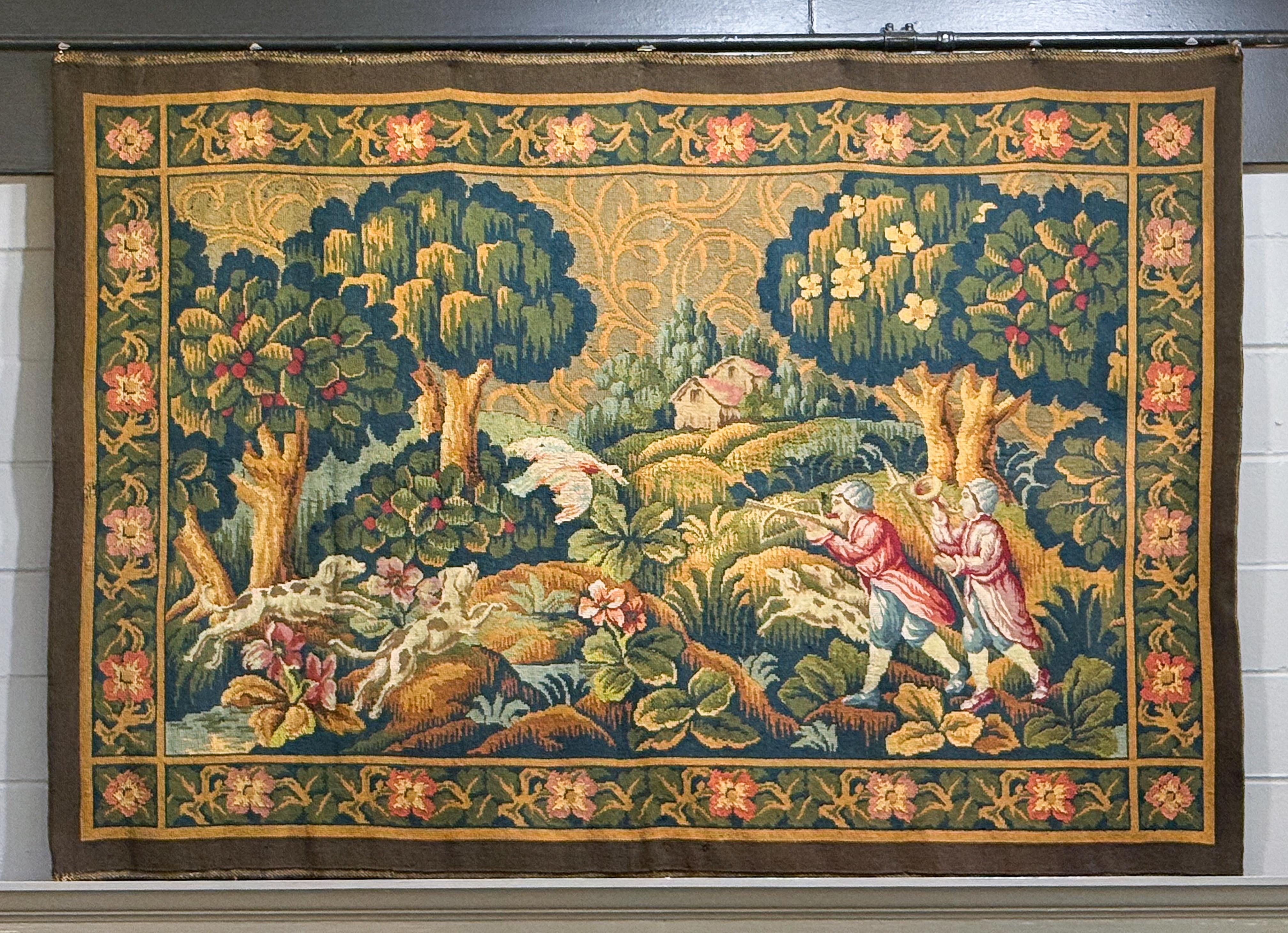 A fine large French Aubusson style tapestry from the 19th century, featuring a scene with two hunters, one aiming a crossbow and another holding a horn, alongside hunting dogs in pursuit of a white bird. A house or chateau is visible in the